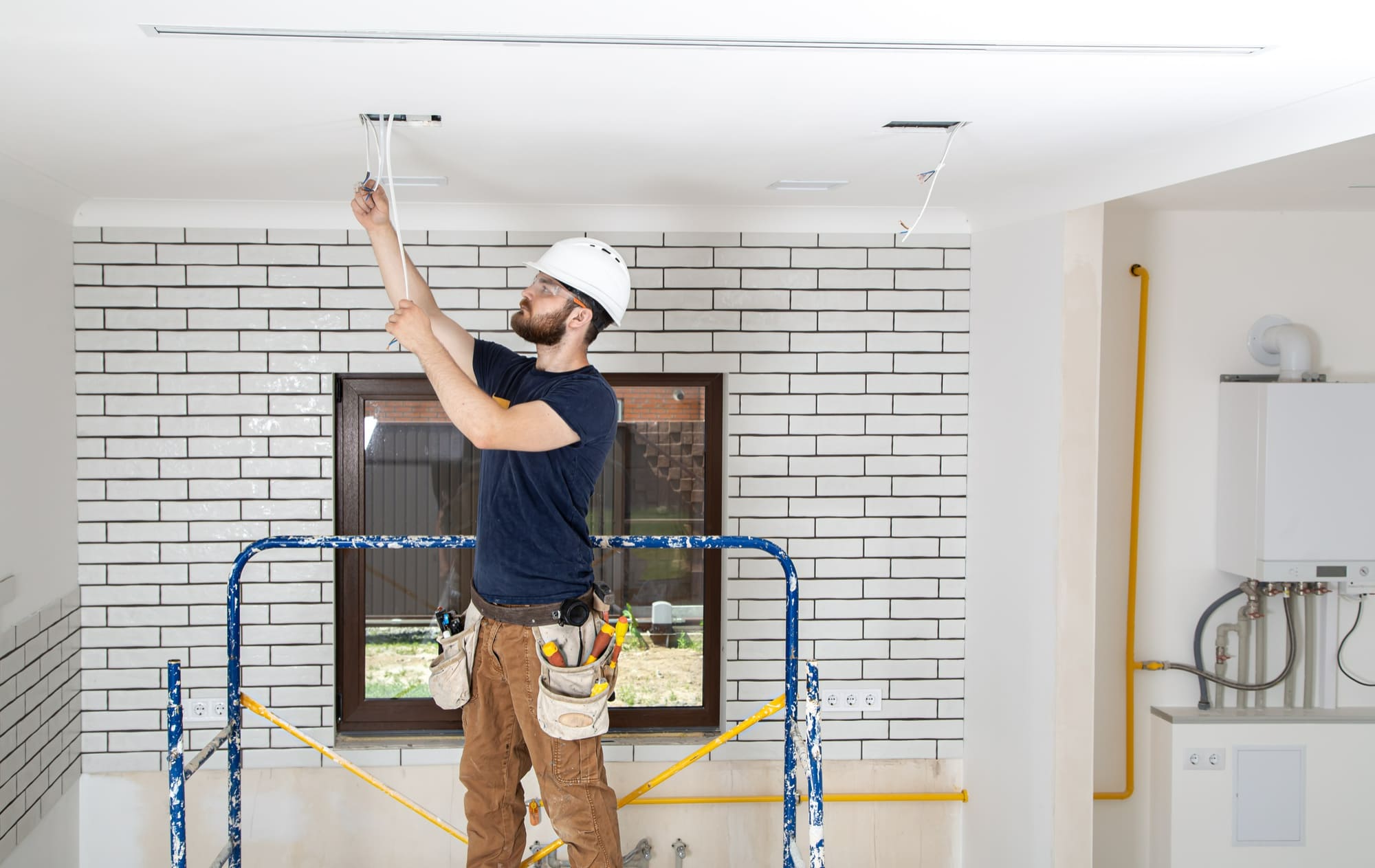 An electrician working on heating and cooling systems in Quebec, standing on scaffolding near a gas furnace and ceiling wiring during a renovation project.