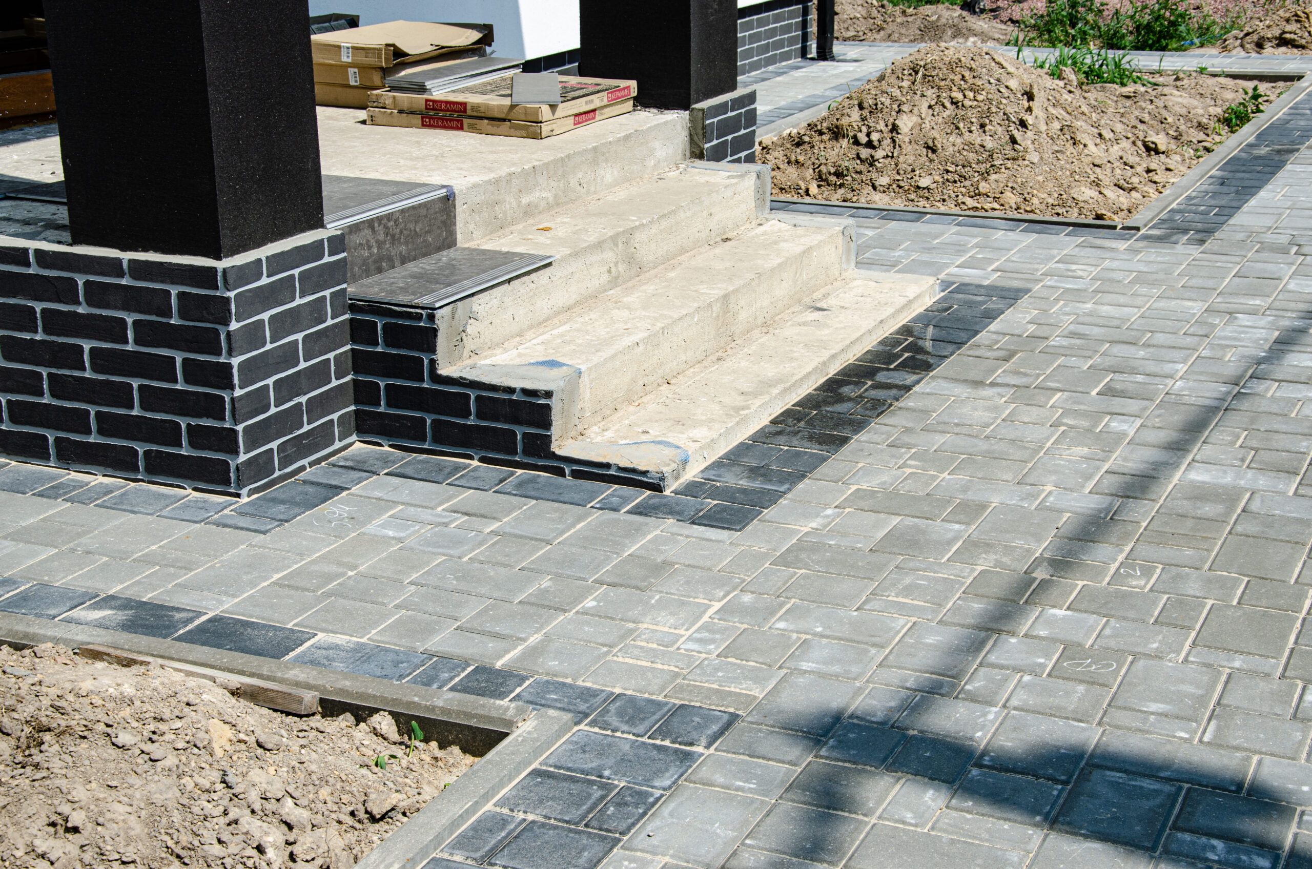 Outdoor construction site in Quebec where paving work is being done, showing a house entrance with black brick steps and a walkway made of gray and dark paving stones.