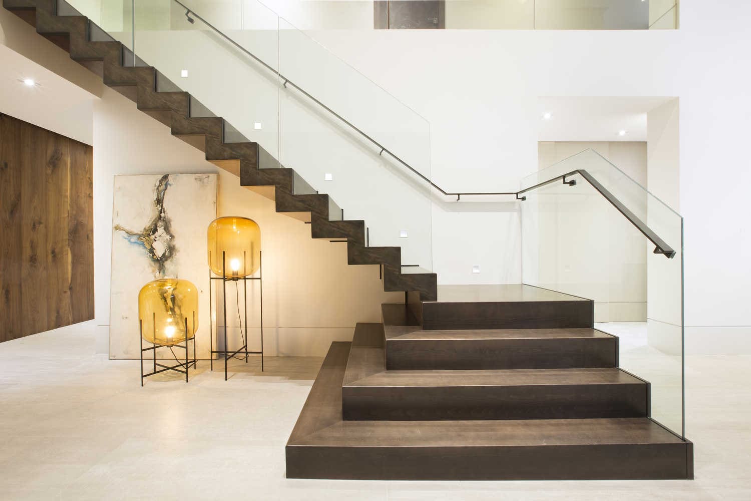 Interior stairs installation Quebec showing a modern staircase with open risers made of dark wood or concrete, featuring a glass railing with a slim metal handrail, two amber-glass floor lamps, an abstract wall painting, and ceiling spotlights for illumination.