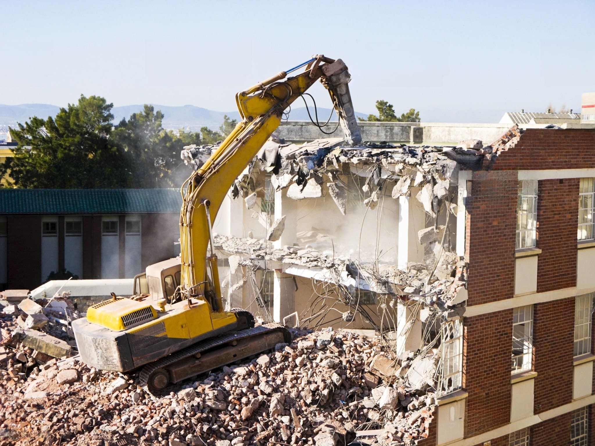 An excavator using a hydraulic breaker to demolish a brick and concrete building, representing professional Demolition Services Quebec and Montreal, with rubble scattered around the construction site.