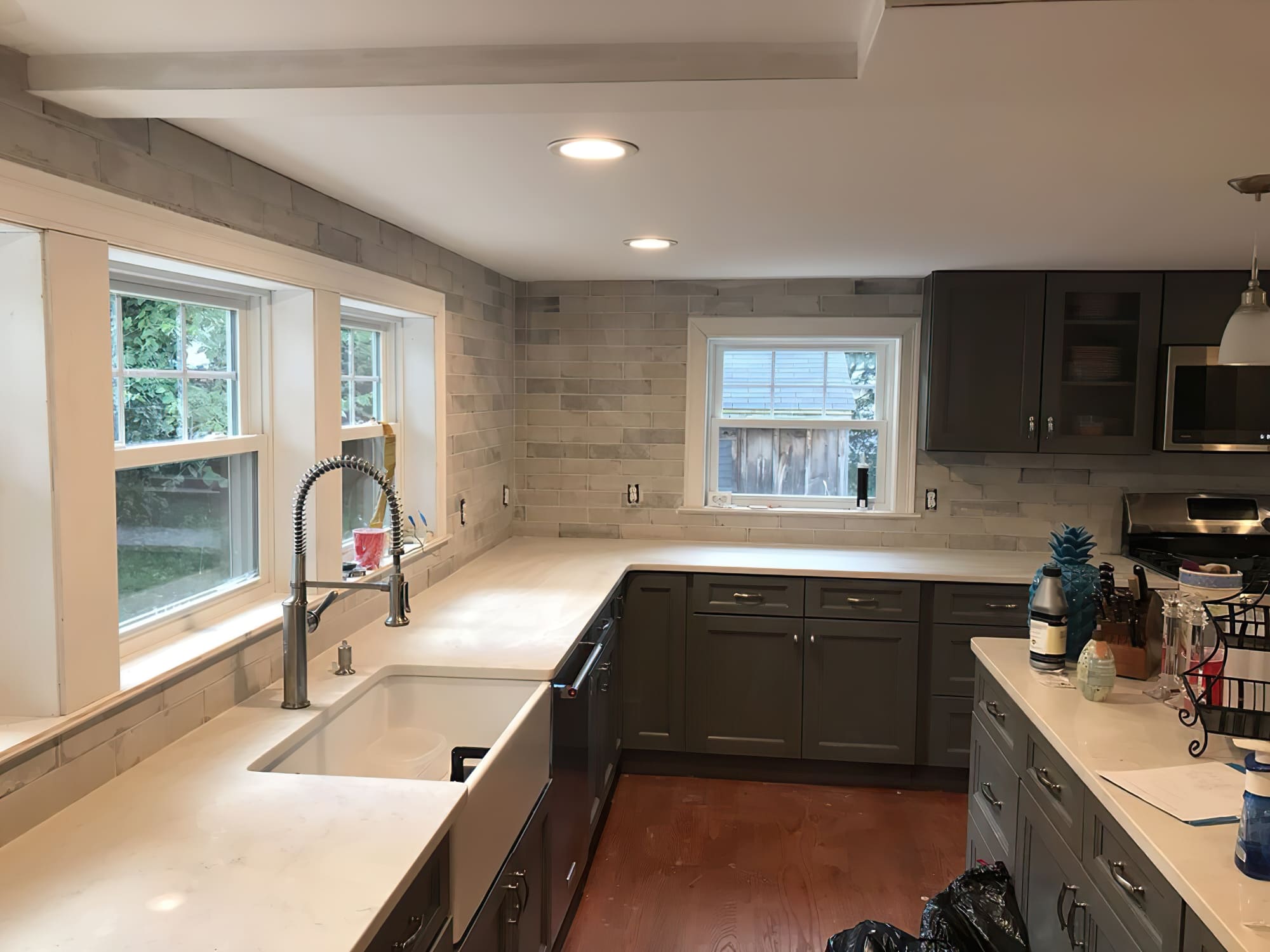 Renovations services Quebec and Montreal featuring a modern renovated kitchen with dark gray cabinets, quartz countertop, stone backsplash, farmhouse sink, and recessed ceiling lights.