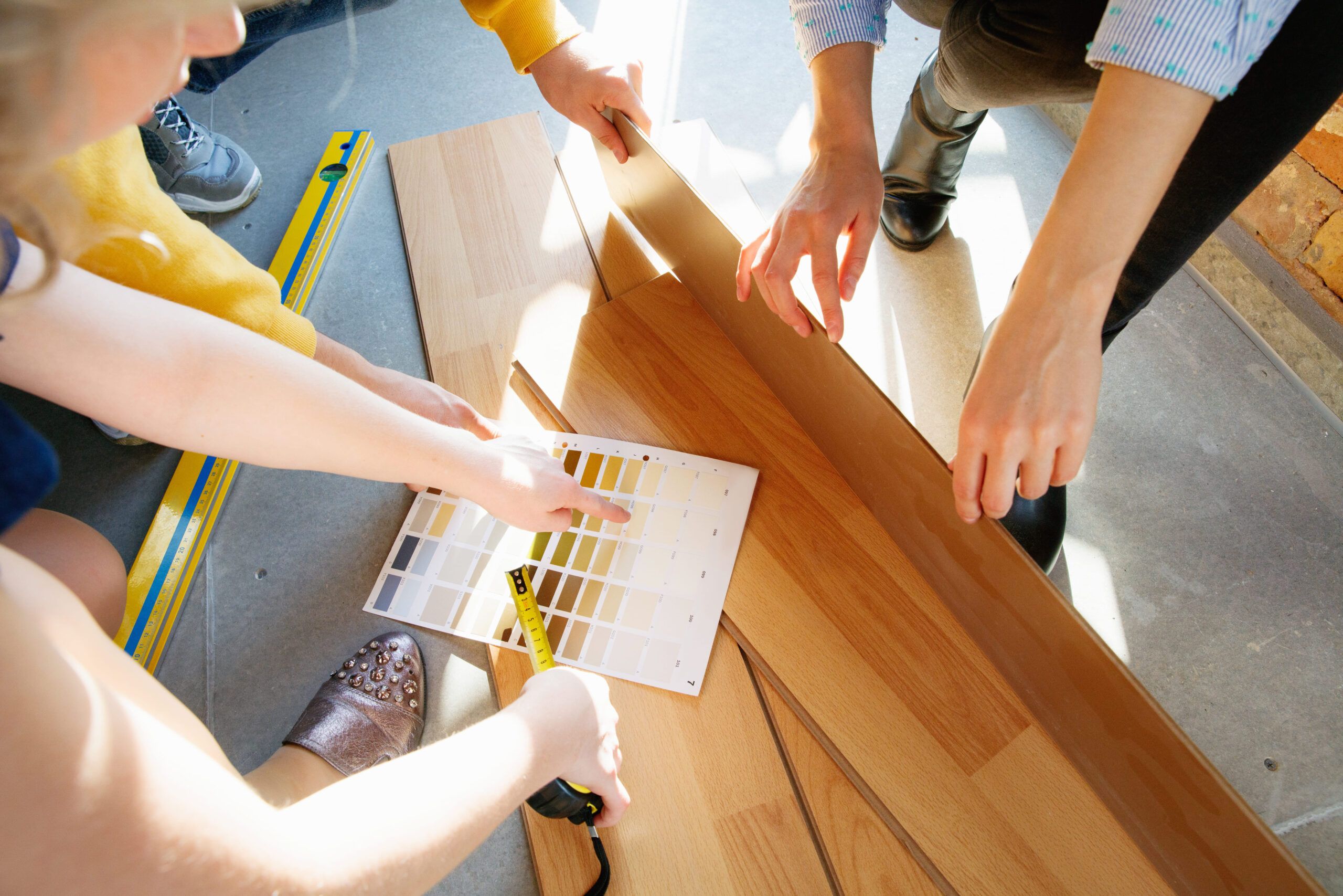 Renovations services Montreal showing a group of people selecting and installing flooring, measuring wooden or laminate planks, and comparing color samples as part of a home renovation or remodeling project.