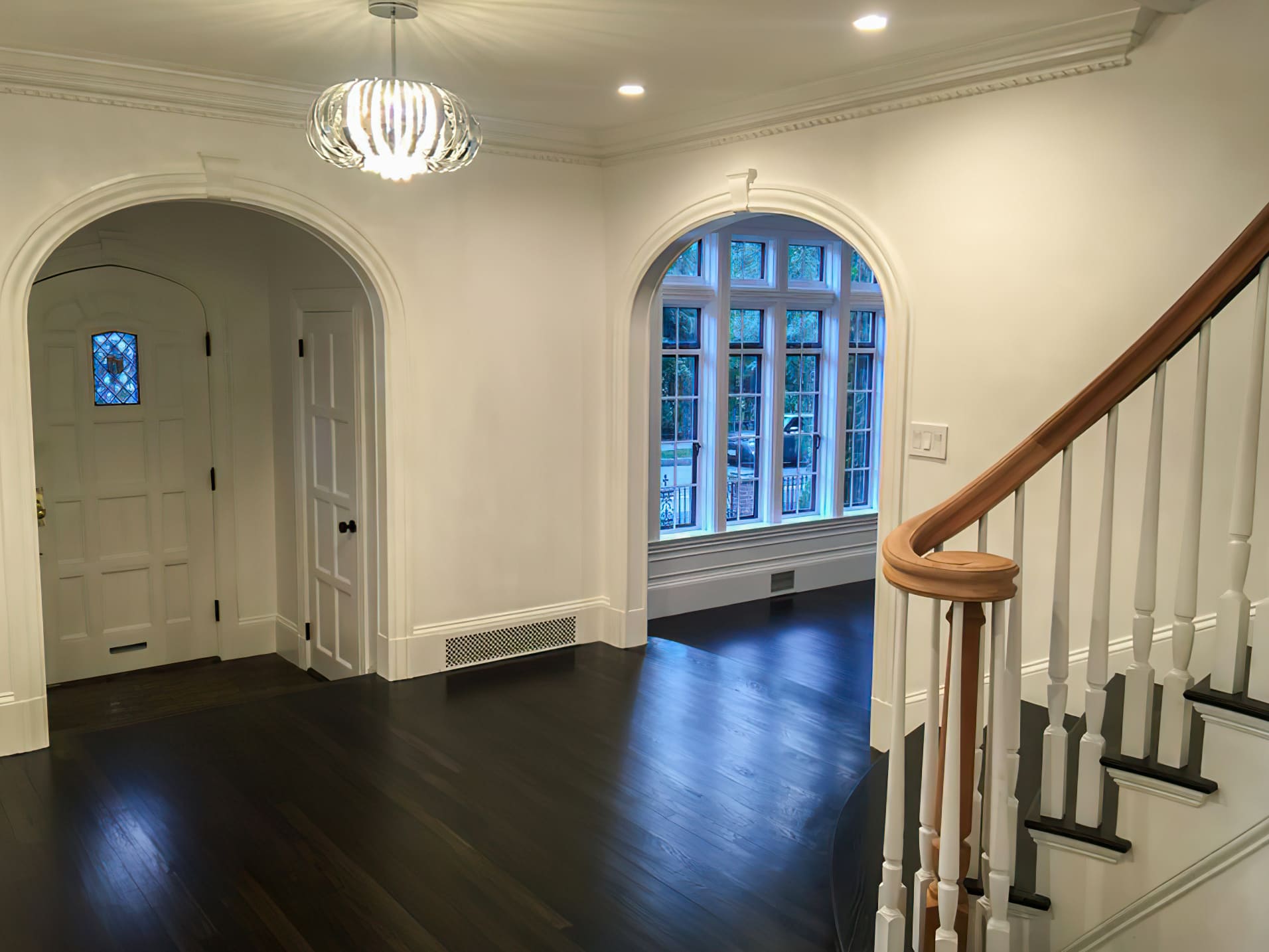 A bright residential entrance hall with dark hardwood floors, white walls, and two large arched doorways. The space features a staircase with a white railing and wooden handrail, a large grid window letting in natural light, and a modern hanging light fixture.