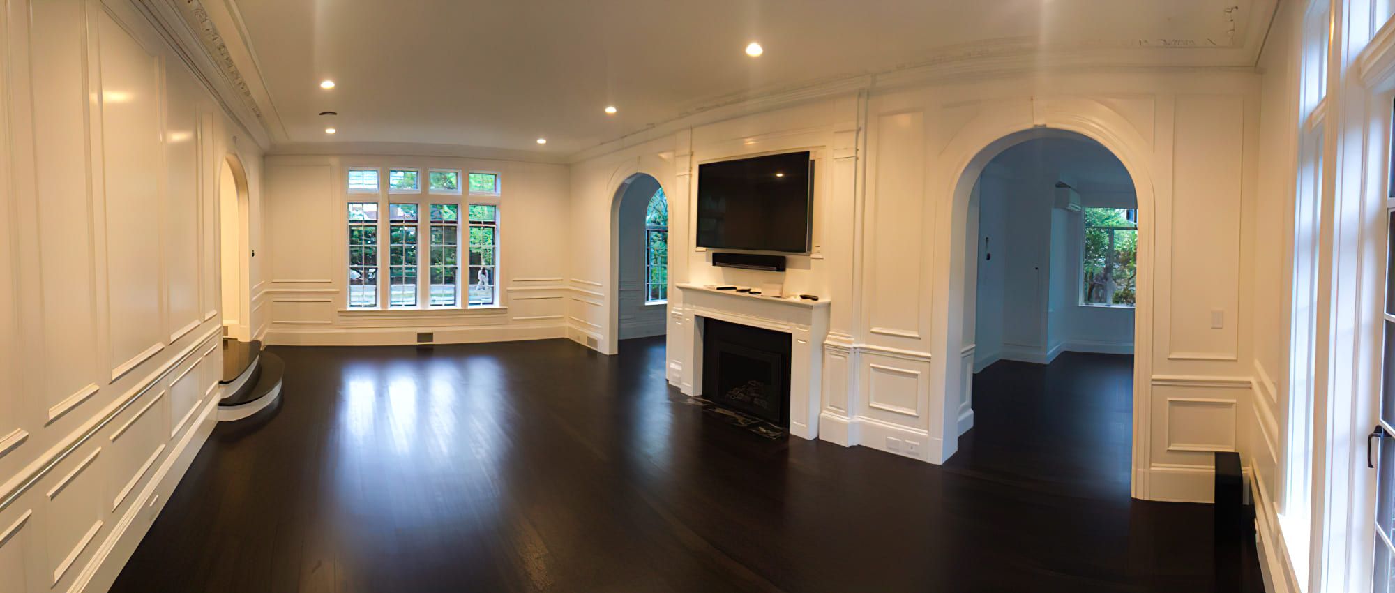 A large empty living room with dark hardwood floors and white walls. The room features a high ceiling with recessed lighting, a fireplace with a wall-mounted TV above it, and several arched doorways leading to other rooms. Large windows, including an arched one, allow natural light to fill the space.
