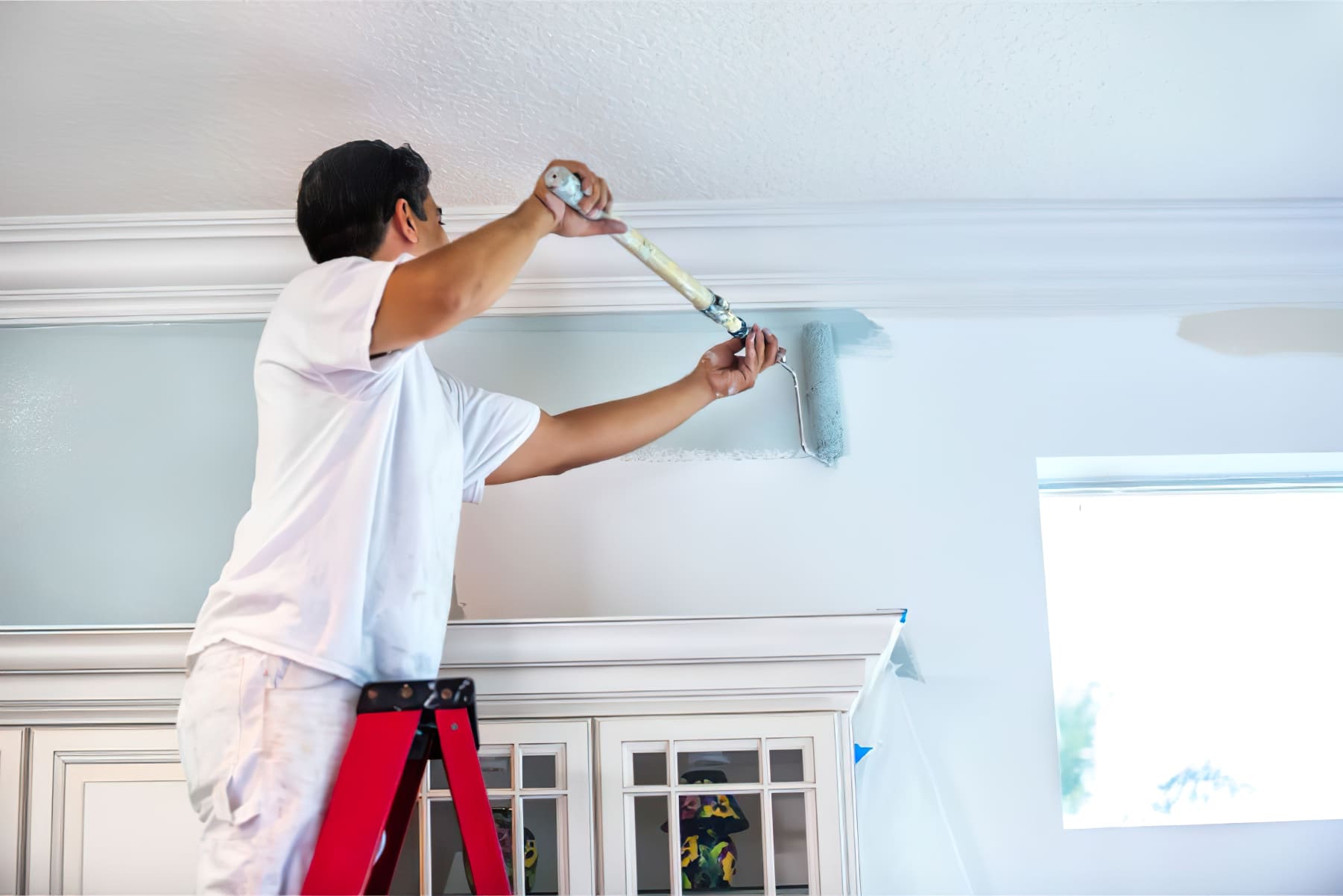 Painting services Quebec showing a painter in white work clothes standing on a red ladder and painting an interior wall light blue with a roller, representing professional residential painting and renovation in Quebec.