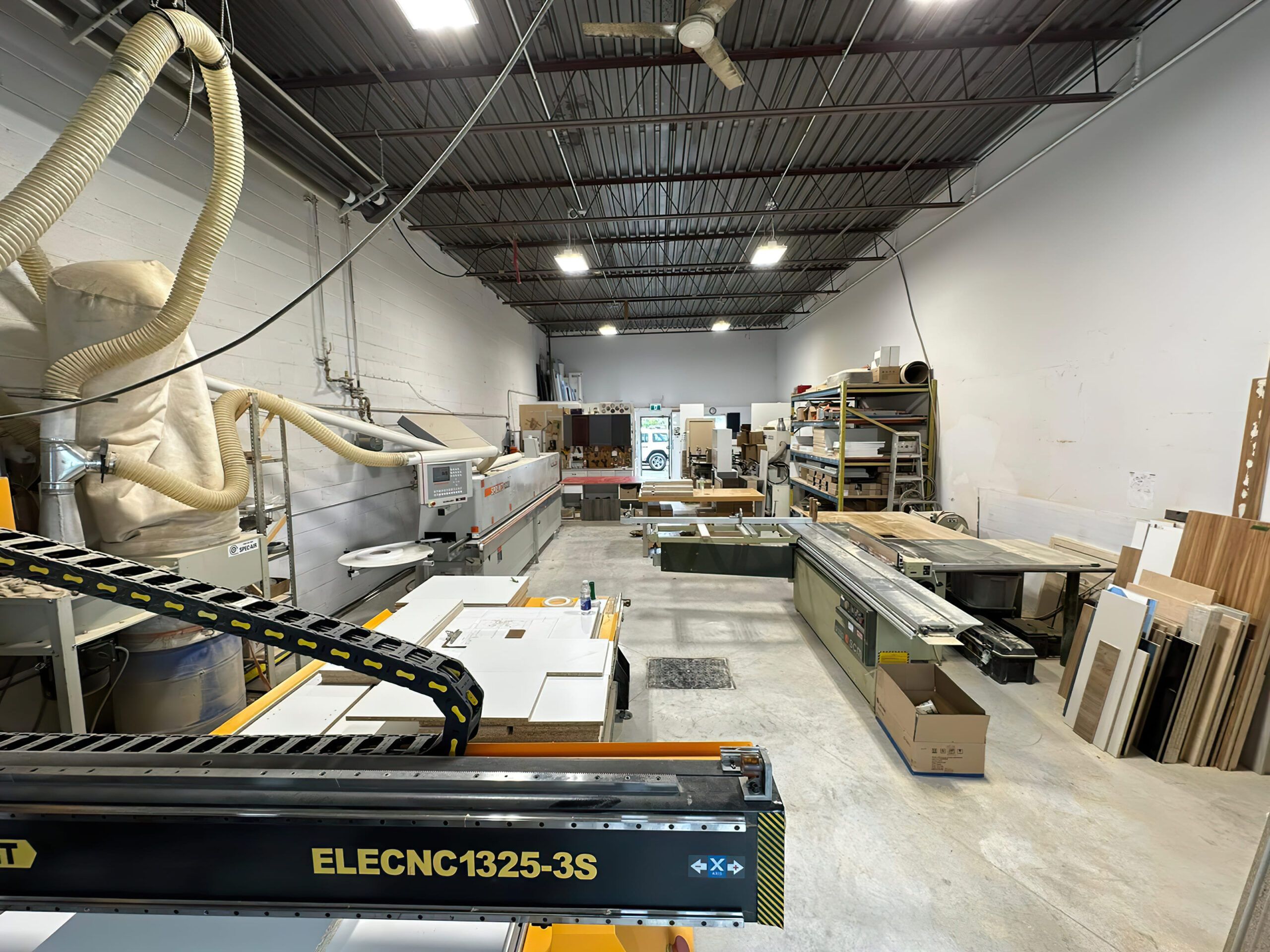 A woodworking and furniture-making workshop in Quebec featuring a CNC machine labeled “ELECNC1325-3S” in the foreground, with dust extraction hoses on the ceiling, lighting fixtures, shelves with materials, and finished wood pieces in the background — representing professional carpentry and joinery craftsmanship.