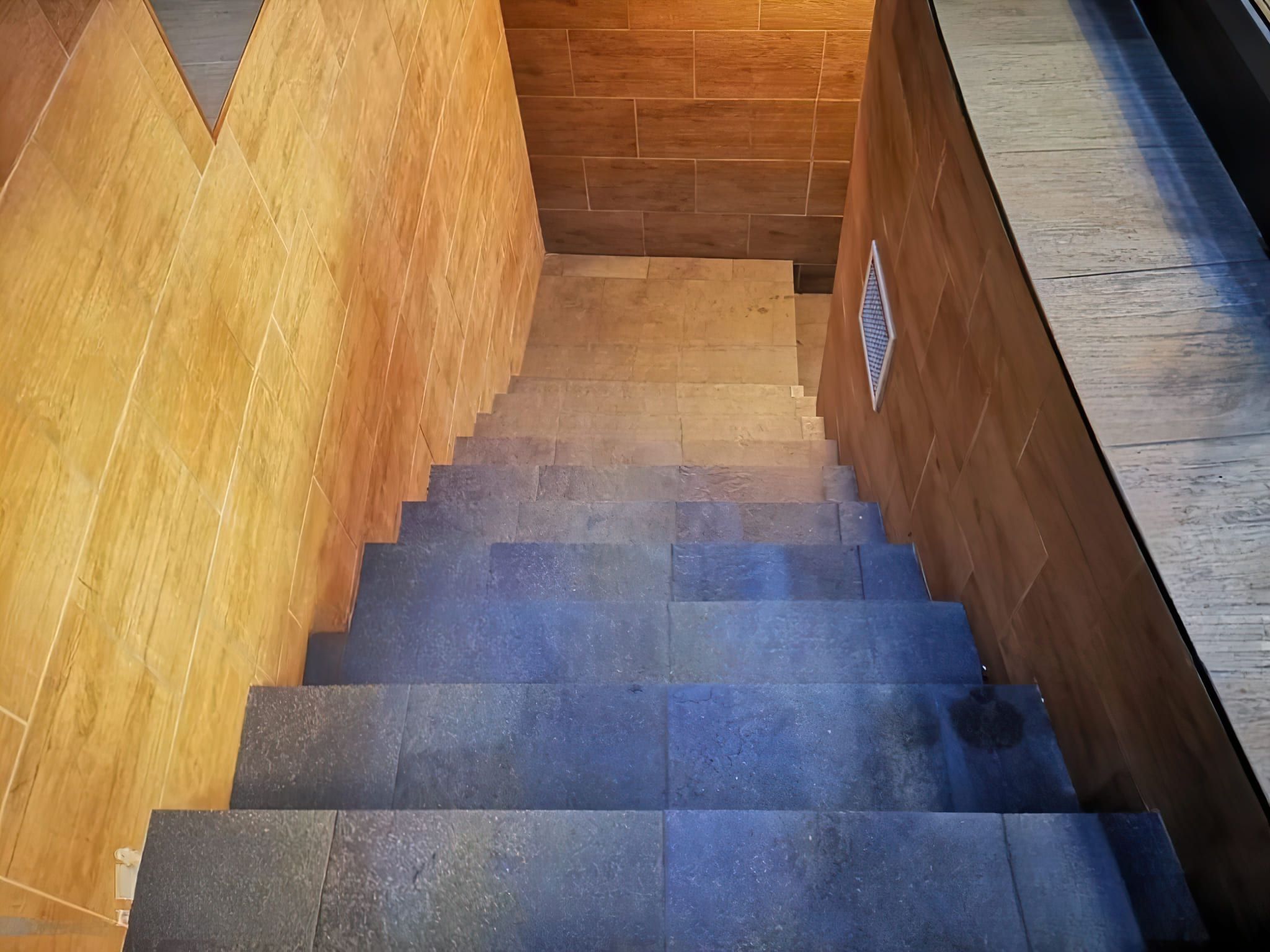 Exterior basement stairs with tiled steps and walls in contrasting dark and light tones, featuring a vent on the right side and a light-tiled landing at the bottom leading to a lower-level entrance.