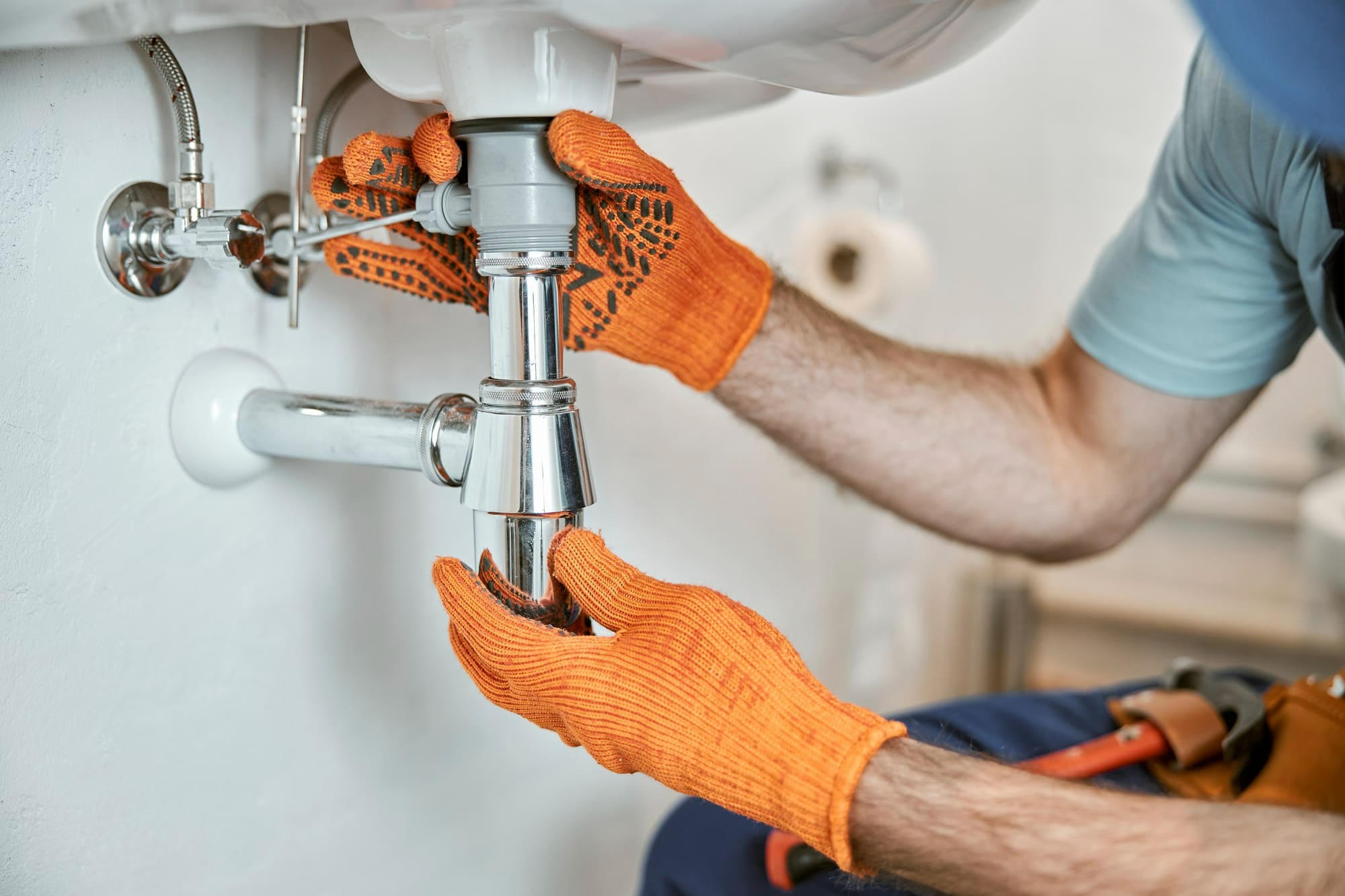 A plumber providing mechanical systems services in Quebec and Montreal, repairing a sink trap (siphon) under a washbasin with tools and wearing orange gloves.