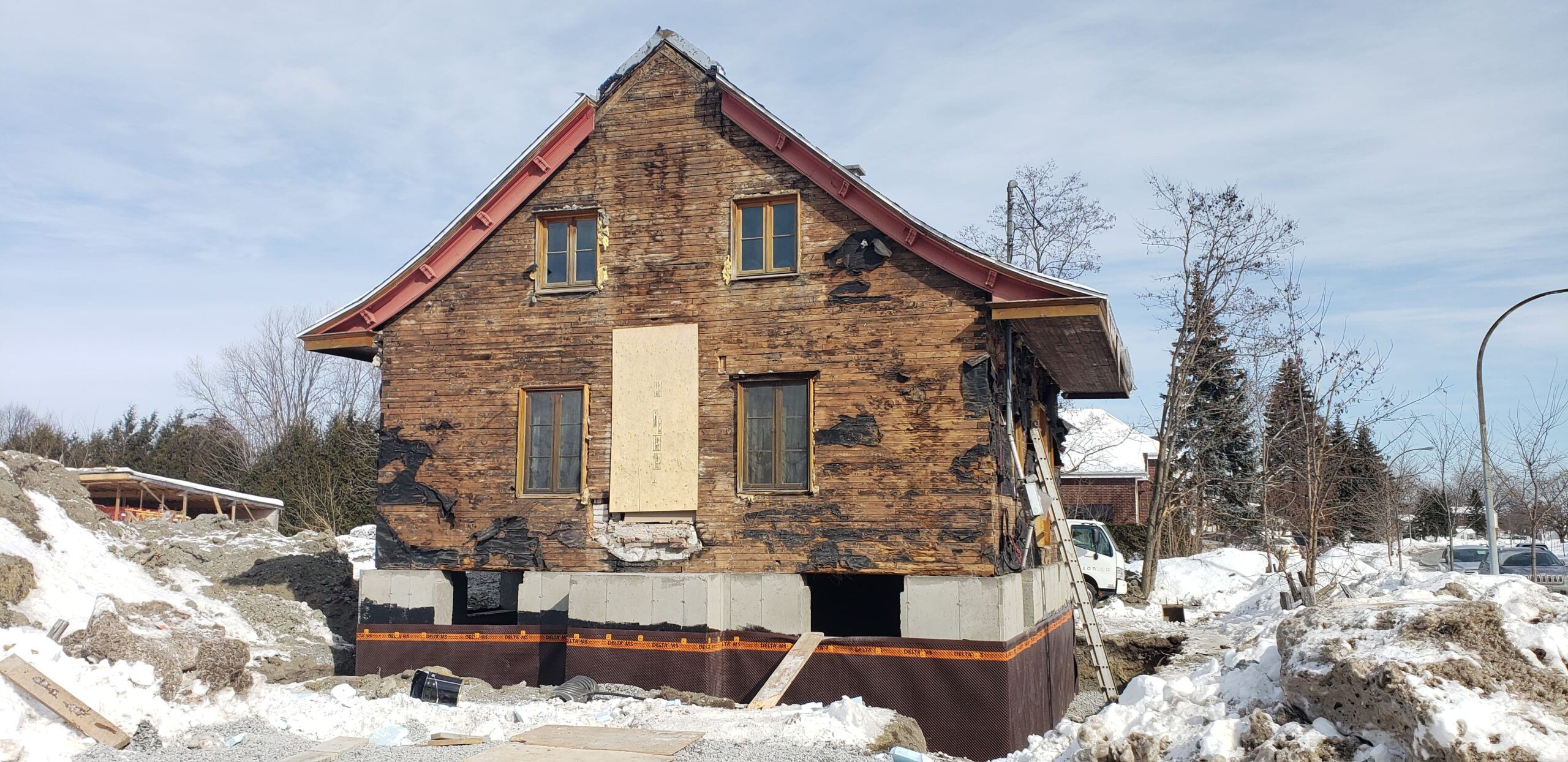 Old detached house under renovation, elevated on a new concrete foundation with exposed wooden framing, surrounded by a snowy construction site with soil piles, scaffolding, and ladders.