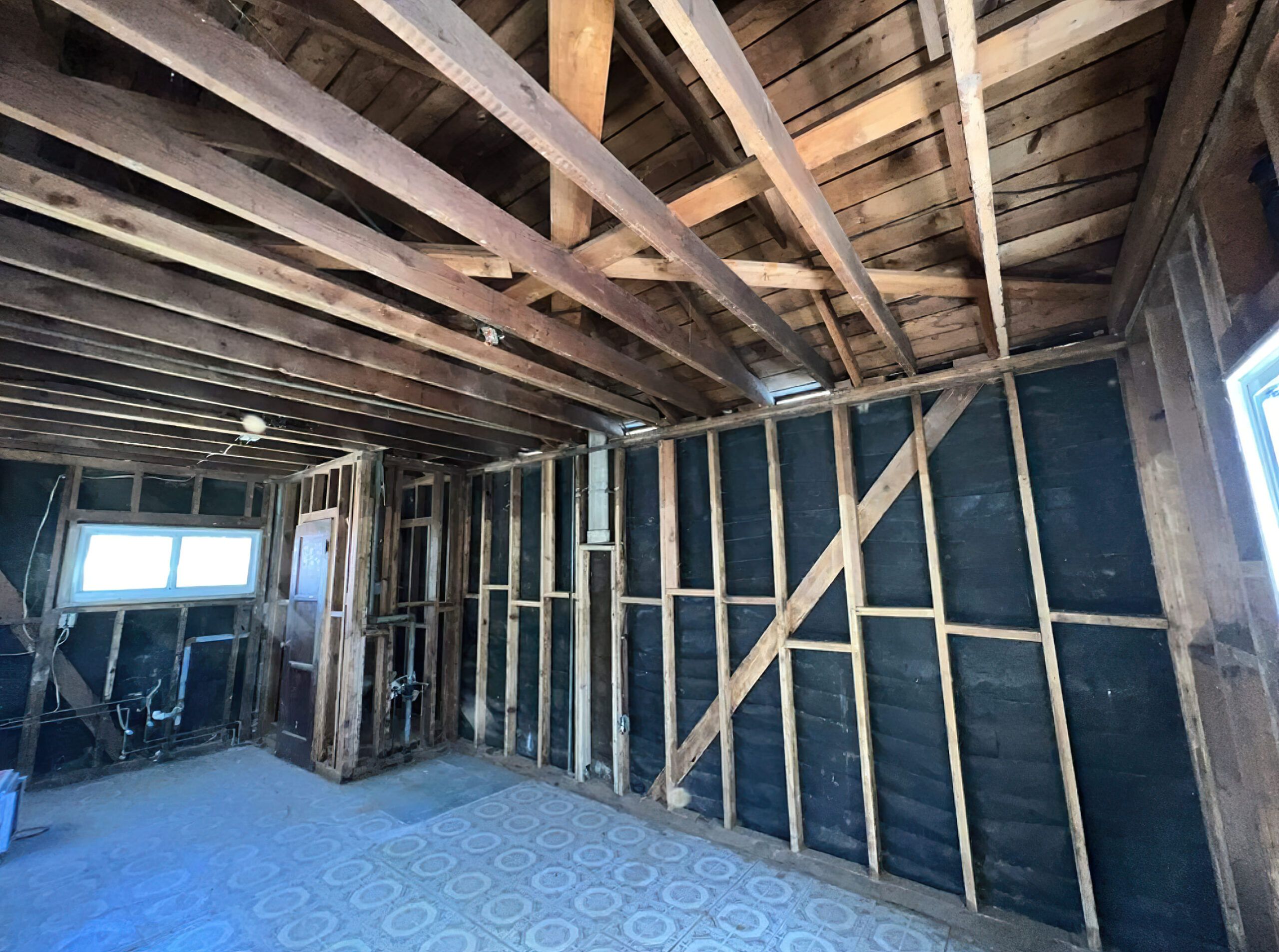 Interior of a building under renovation in Quebec showing exposed wooden ceiling beams and wall studs, black vapor barrier or insulation on walls, and a tiled floor with a circular pattern.