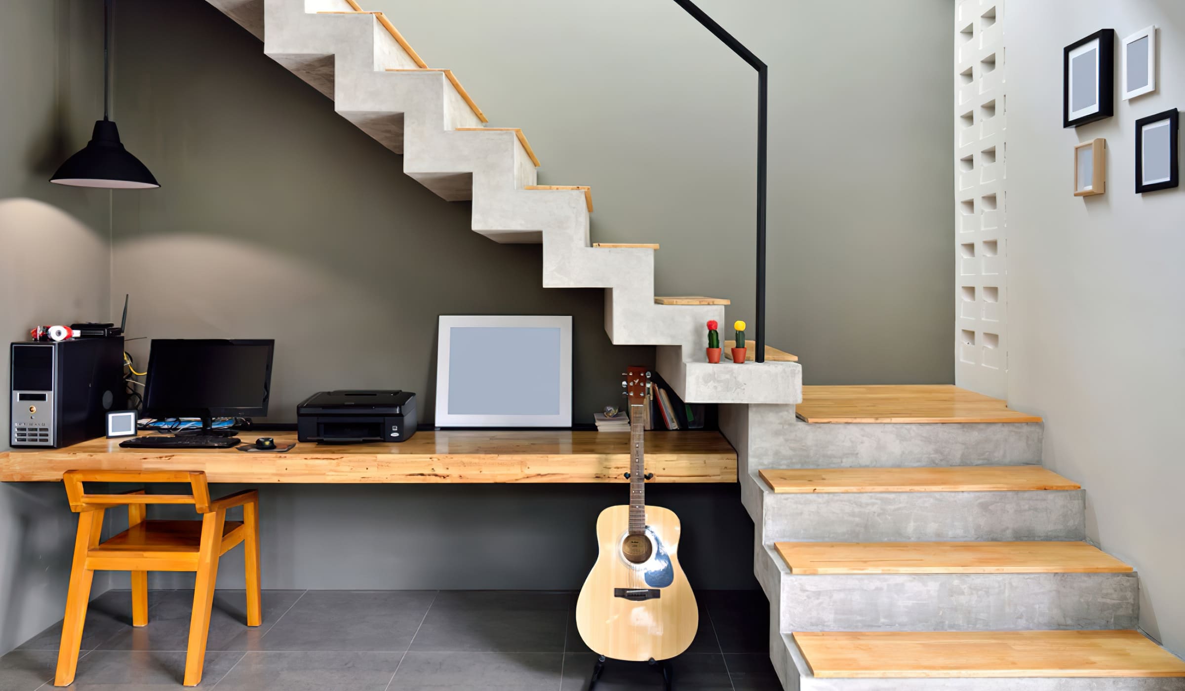 Interior stairs installation Quebec featuring a modern concrete and wood staircase with open risers, serving as both a room divider and workspace; the area beneath includes a wooden desk, computer, and guitar, showcasing a minimalist and space-saving interior design.