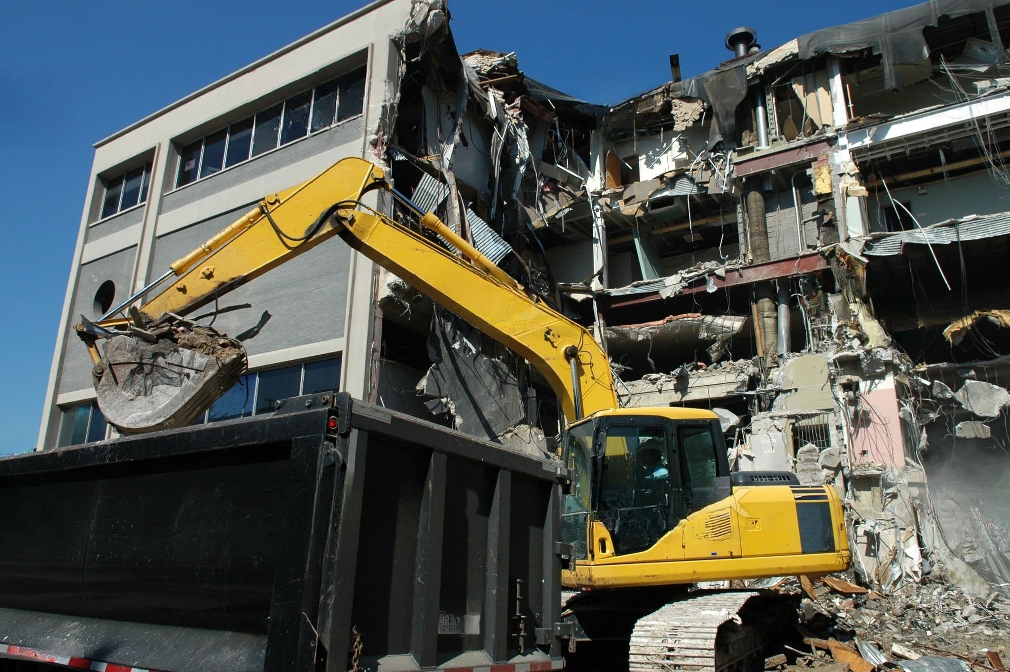 Commercial Demolition Quebec showing an excavator loading debris into a truck during the demolition of a partially destroyed building.