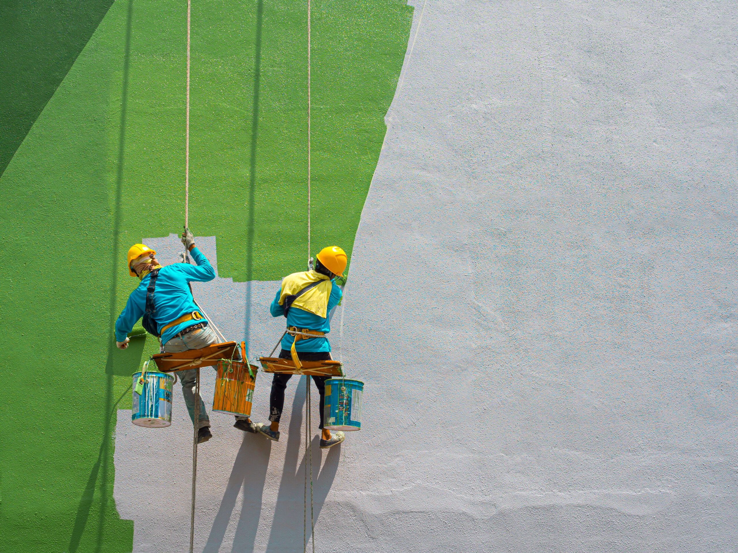 Two painters suspended on a high-rise building facade applying exterior paint. They are equipped with safety harnesses and helmets while performing professional facade painting work.
