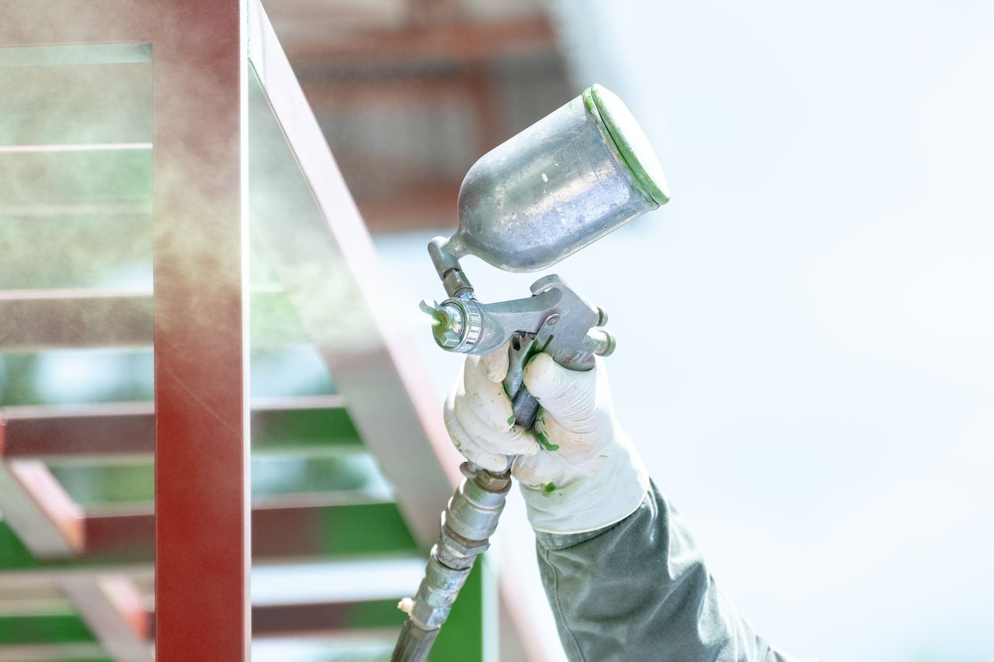 Worker spraying red paint onto a metal structure using a spray gun during an exterior painting project in Quebec. The worker is wearing protective gloves and work clothes for safety.