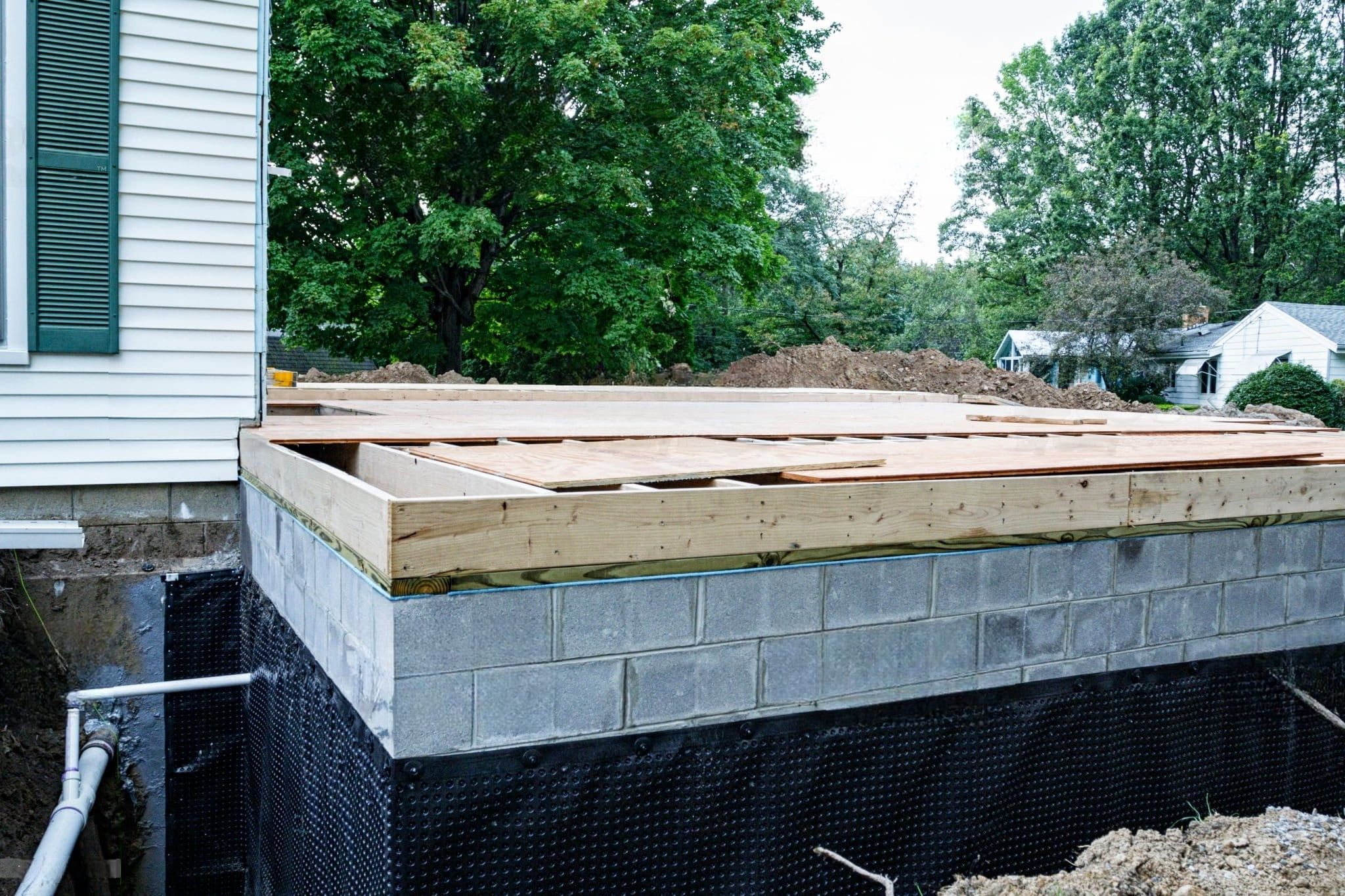 Waterproofing & French Drain Services Quebec showing a house foundation with concrete blocks, a black dimpled membrane for moisture protection, and a gravel layer for drainage and frost prevention.