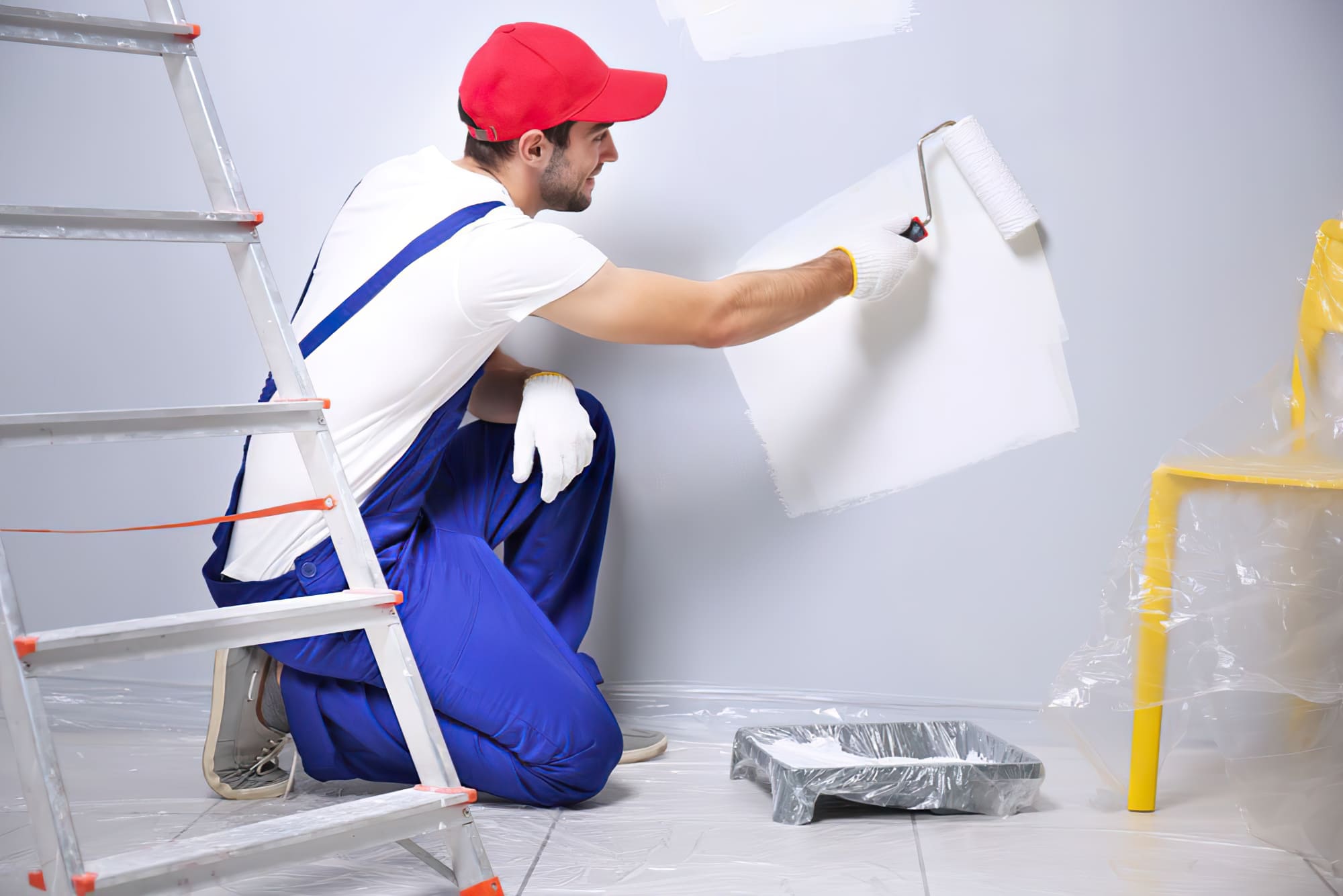 Painting services Quebec showing a painter in a blue overall and white cap kneeling while painting a wall with a roller. The scene represents professional residential painting and renovation work in Quebec.