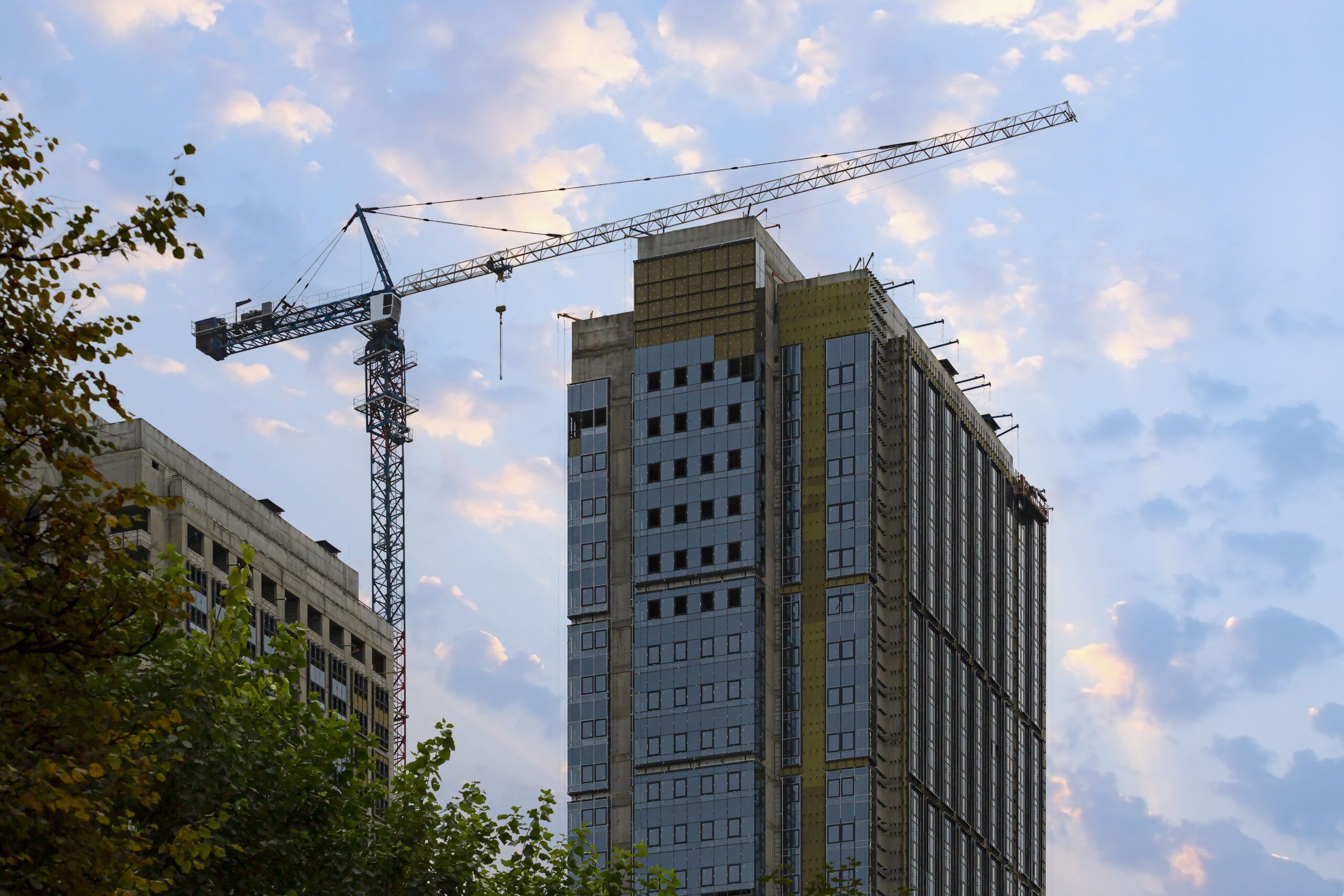 New Construction Quebec showing a city construction site at sunset with a tower crane beside a high-rise building under development partially covered with insulation material and urban buildings in the background.