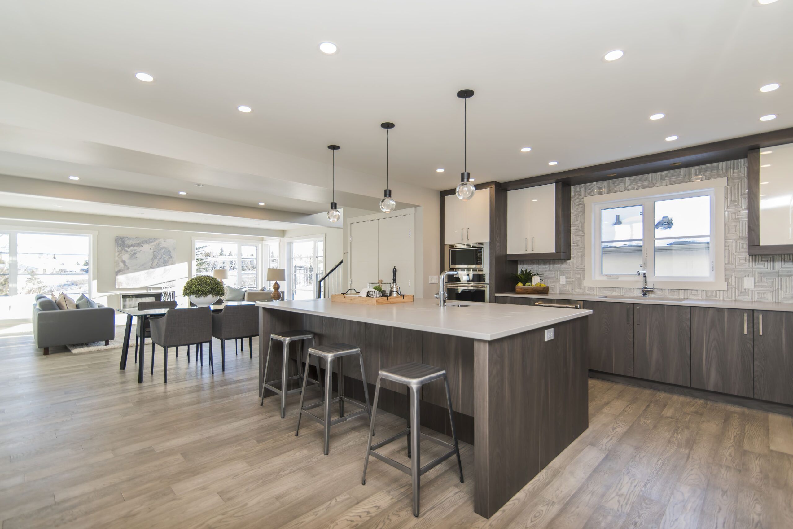 Renovations Services Quebec featuring a modern open-concept kitchen with a large island, white and dark wood cabinets, light wood flooring, pendant lights, and an adjoining dining area with a dark table and gray chairs.