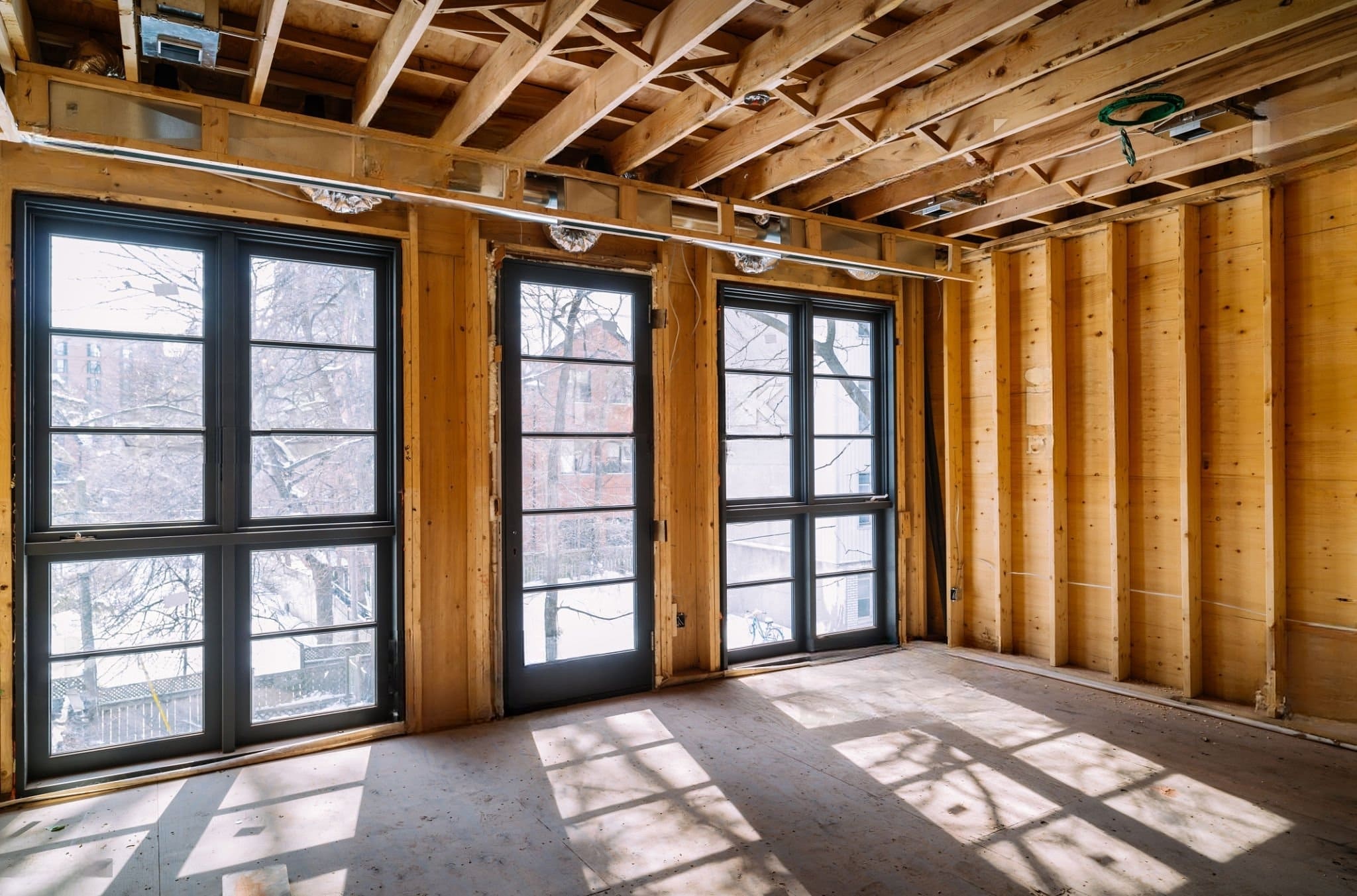 Windows & Doors installation Montreal — A house under construction with wooden wall and ceiling frames, large black-framed window and door openings, and an unfinished concrete floor allowing natural light into the space.