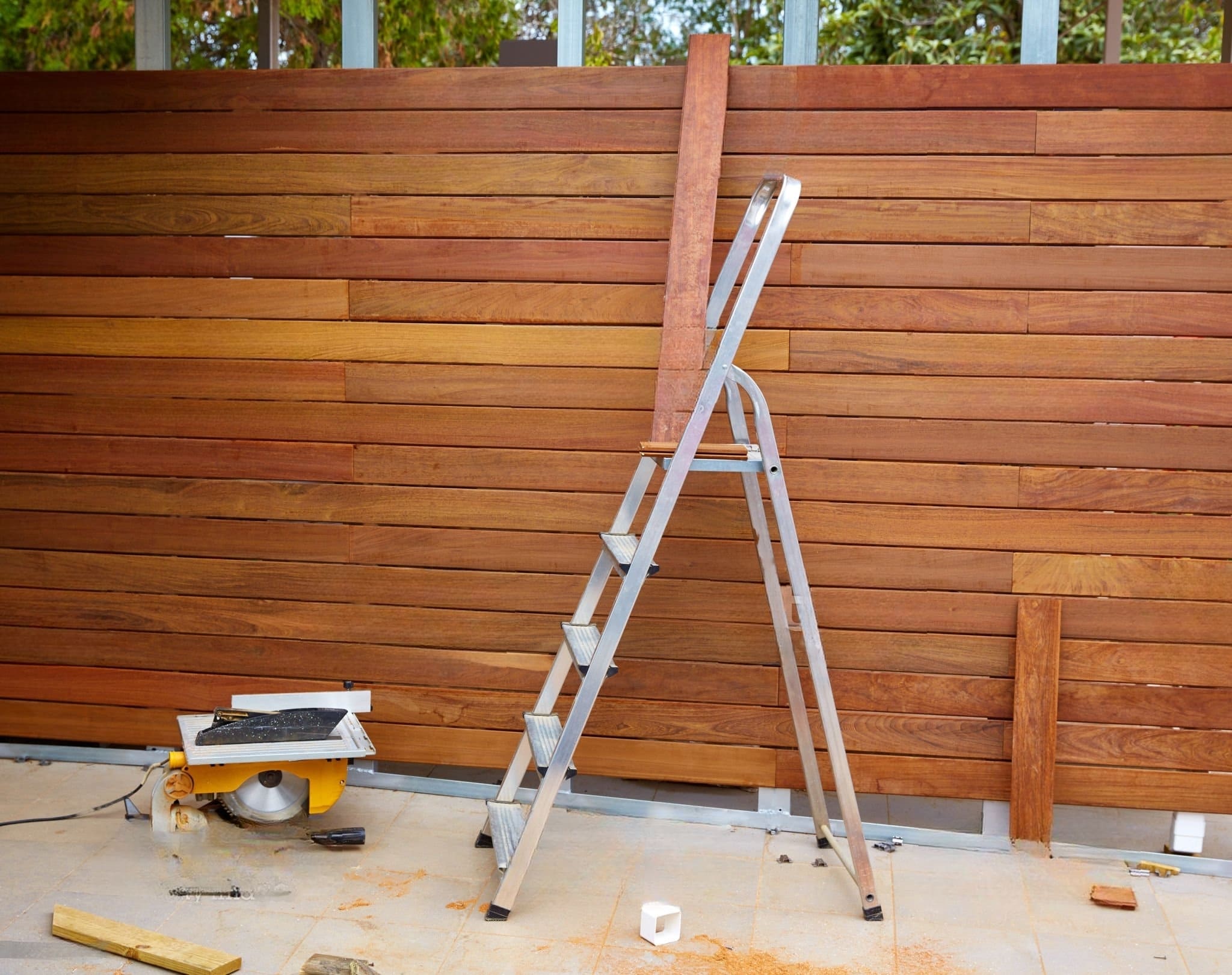 A construction site showing a partially built wooden deck and fence as part of a Decks & Fences Solution in Quebec, with a ladder, circular saw, and tools beside reddish-brown treated wood planks
