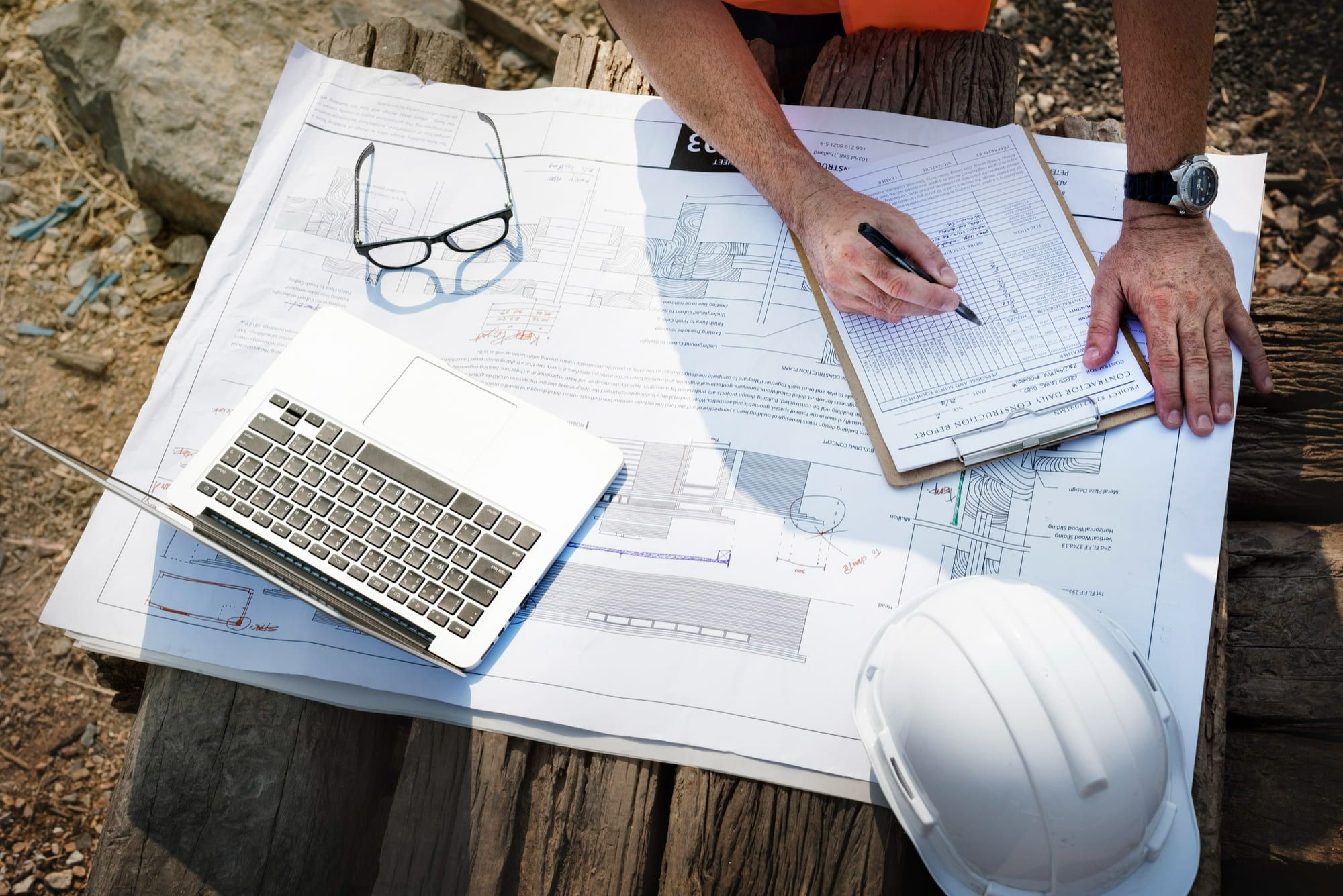 Architecture and engineering services Quebec and Montreal showing contractor reviewing construction blueprints and daily site report with laptop safety helmet and clipboard on desk.