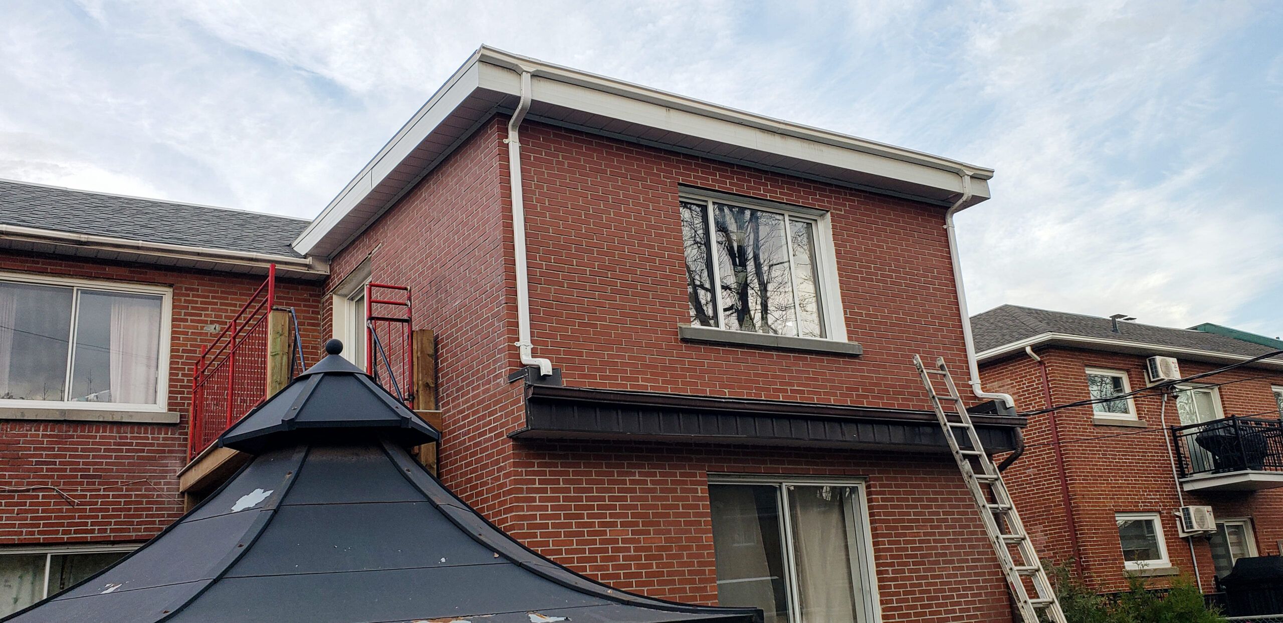 Home addition project in Quebec showing the side of a red brick house with white gutters and a ladder against the wall, suggesting renovation or maintenance work, with part of a pavilion roof visible in the foreground.