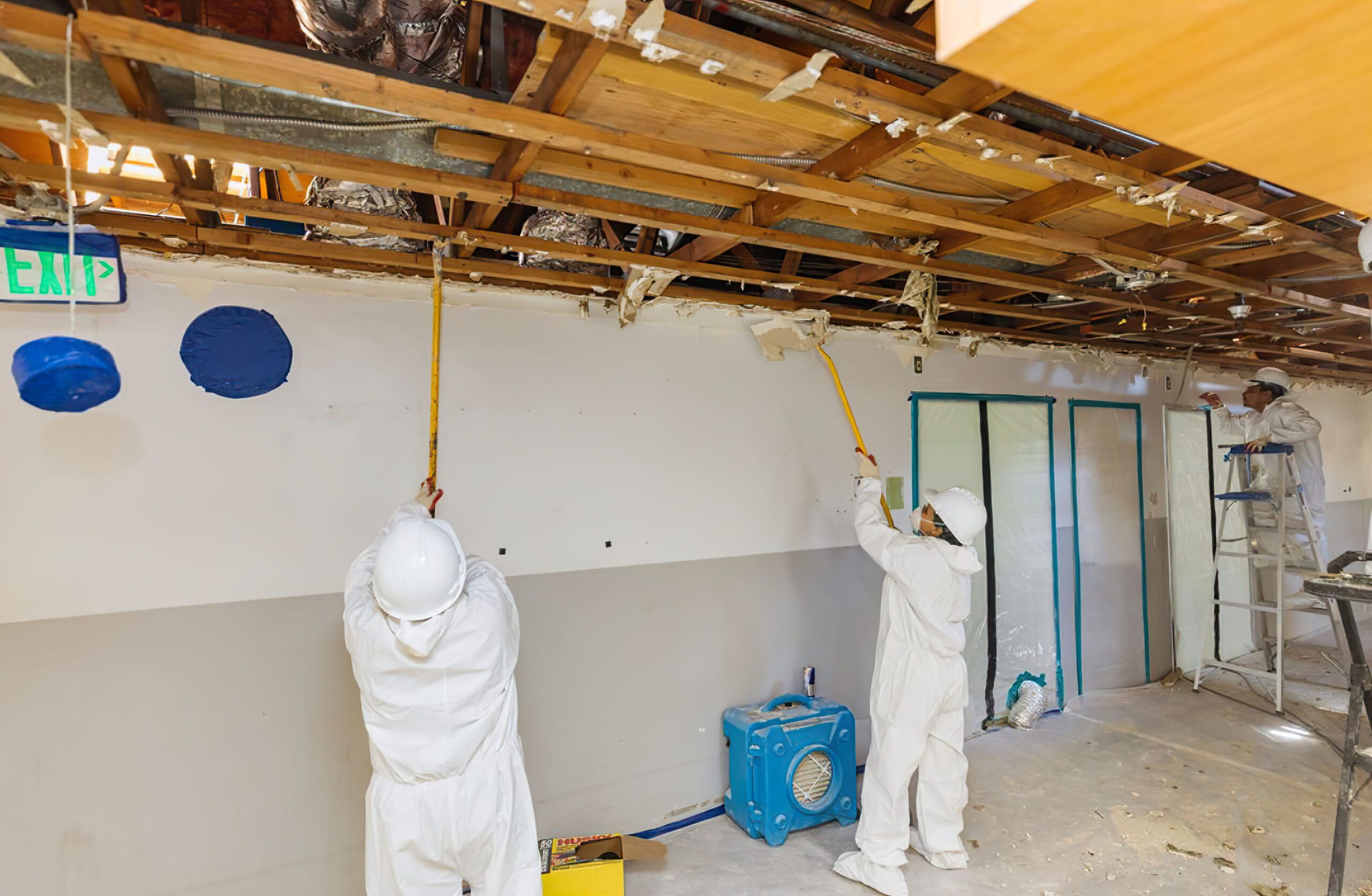 workers in protective suits renovating a room in Quebec removing old ceiling materials with long tools exposing wooden beams during early stage of decontamination or restoration work.