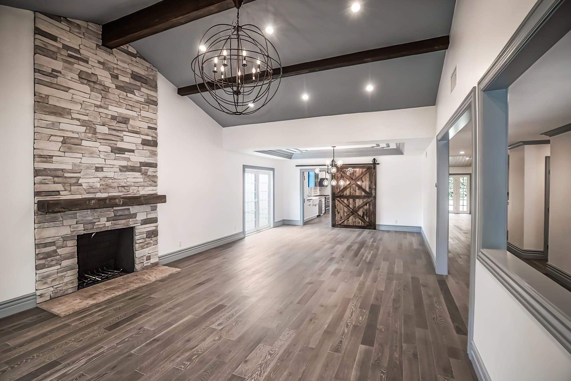 House remodeling Quebec showcasing a Mountain Craftsman style interior with natural wood flooring, vaulted ceiling, stone fireplace, wooden barn door, and a modern spherical chandelier.