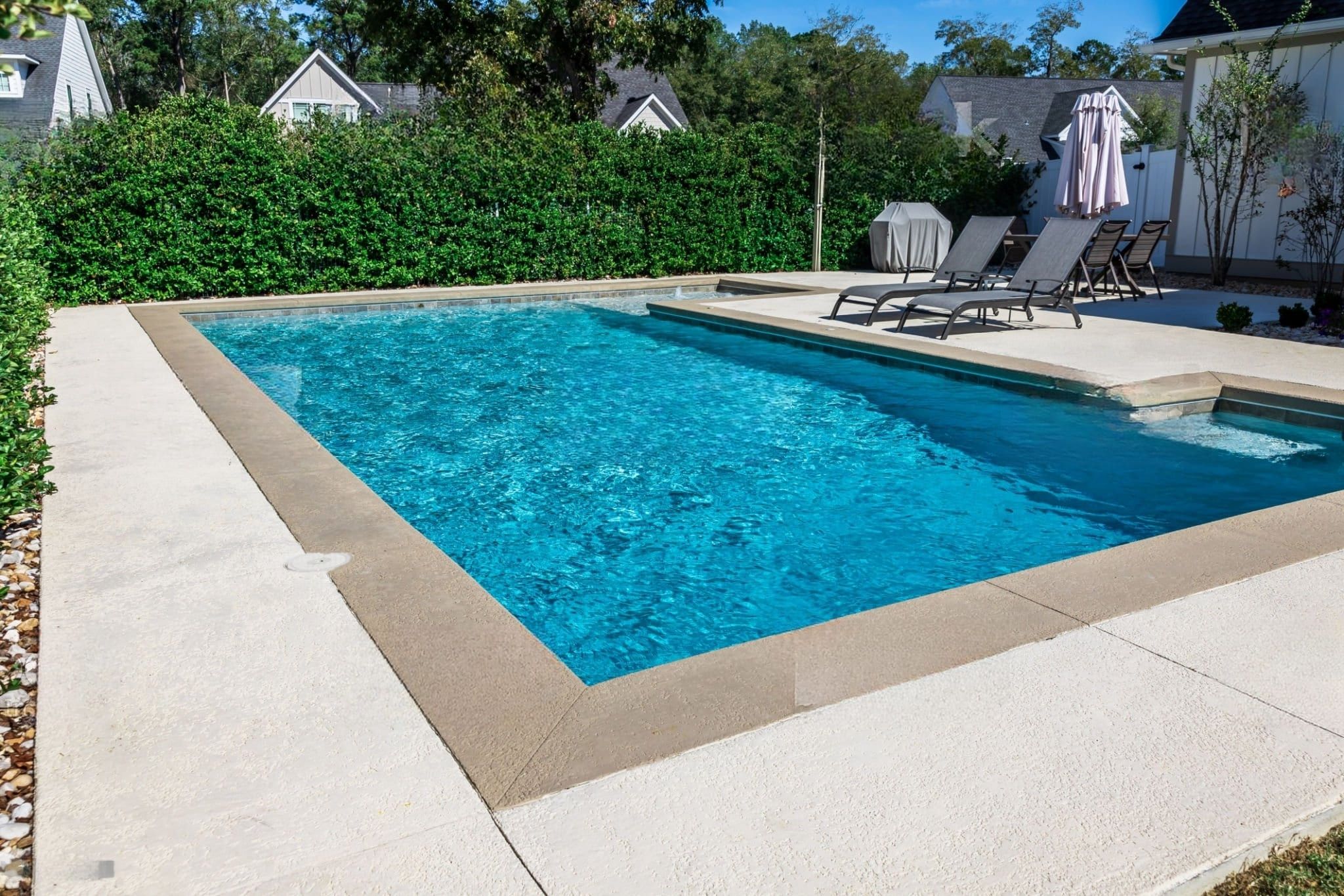 custom pool builders Quebec — modern rectangular backyard swimming pool with light stone decking, lounge chairs, a grill, and a sun umbrella surrounded by green hedges.