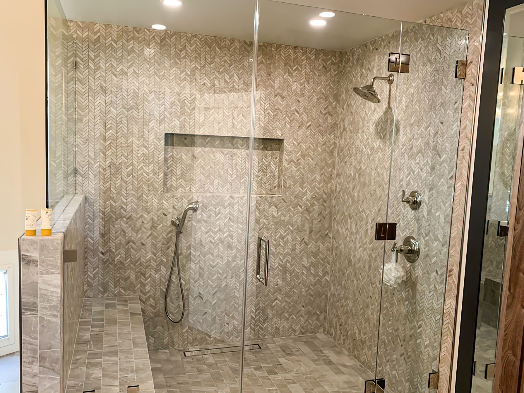 Modern walk-in shower with chevron-patterned tiles, built-in bench, glass enclosure, and sleek metal fixtures, showcasing elegant bathroom design and remodeling in Quebec for contemporary homes.