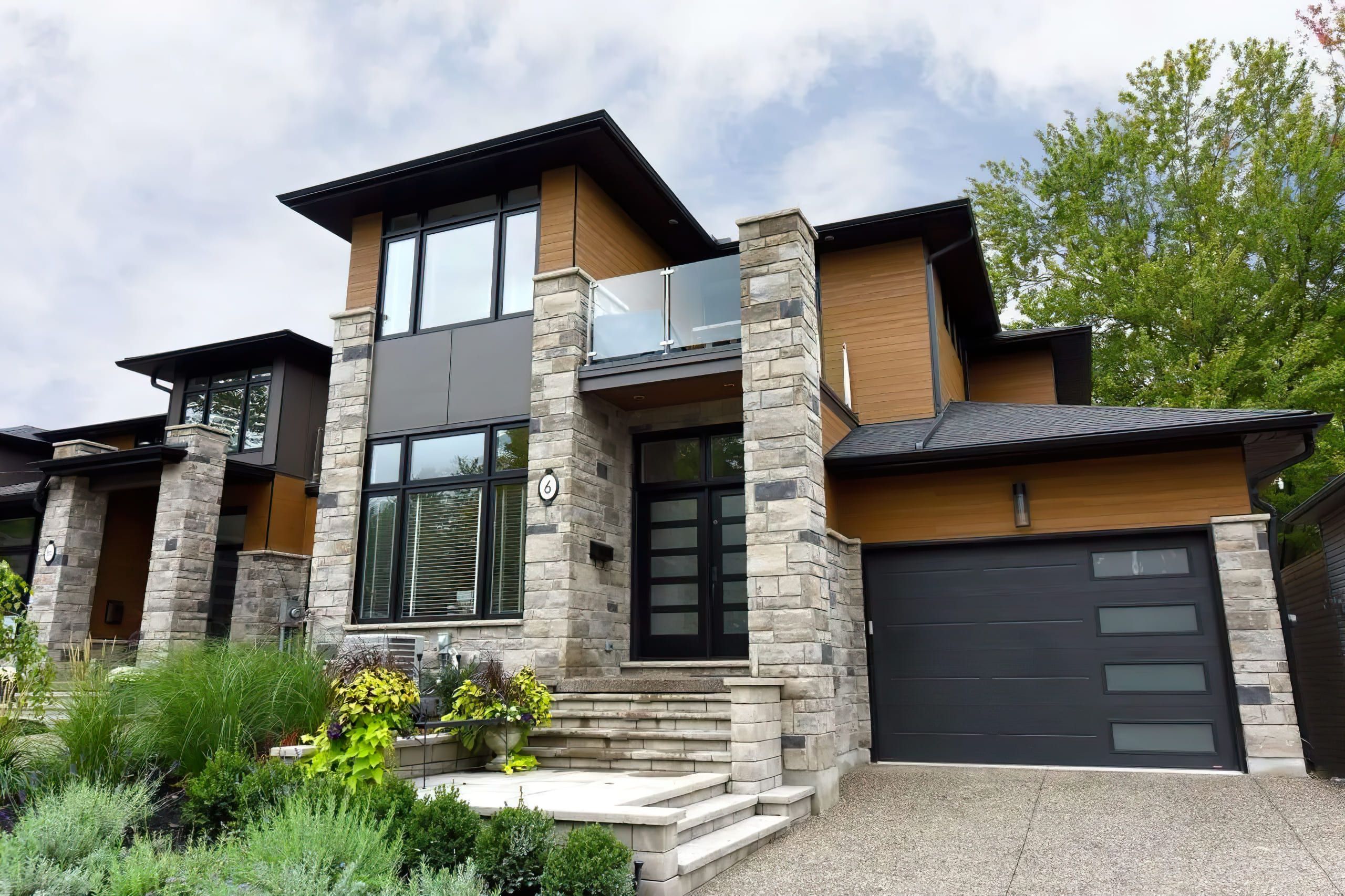 modern residential construction Quebec featuring a two-story house with stone, wood, and gray panel façade, black window frames, glass balcony, and contemporary architectural design.