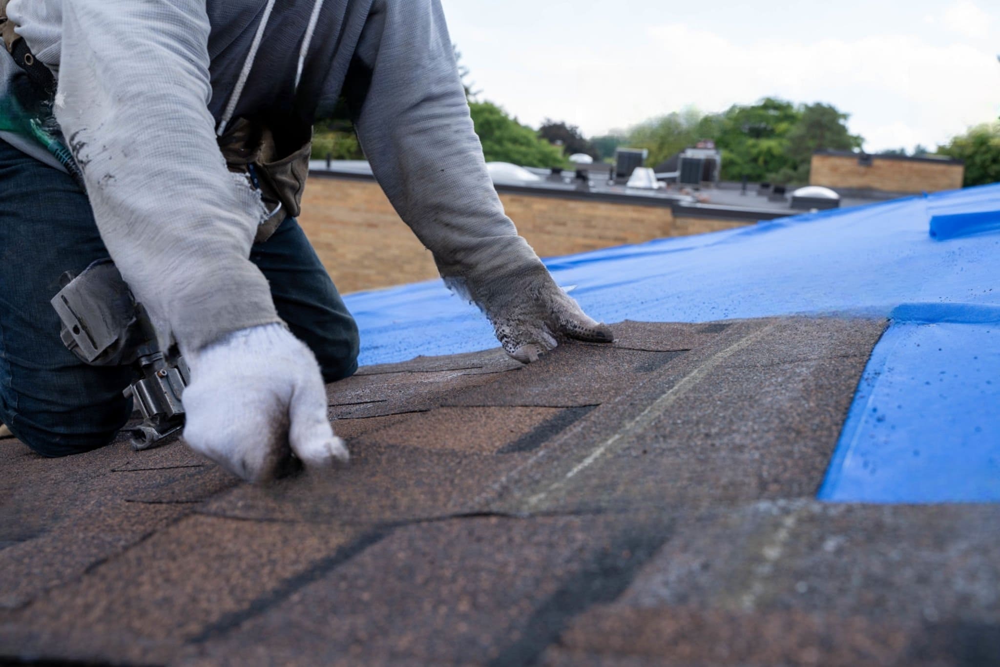 A roofer in Quebec installs brown asphalt shingles over a blue waterproof underlayment during a roof installation project. The worker is wearing gloves and a safety harness while carefully overlapping the shingles to ensure proper sealing and moisture protection.