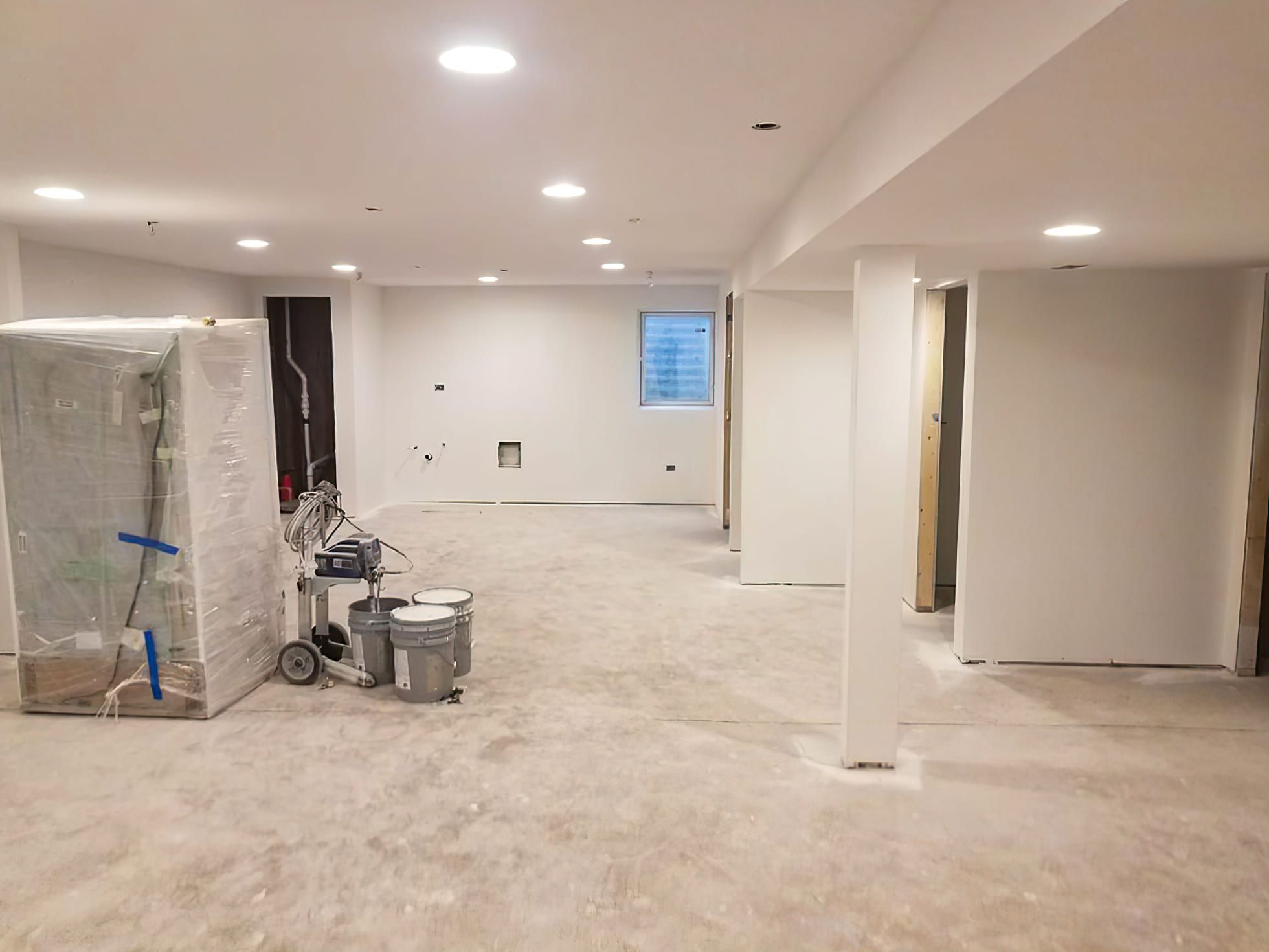 A basement renovation in Montreal showing freshly painted white walls and a ceiling with recessed lights, with painting tools and paint cans on the floor — illustrating ongoing basement finishing services.