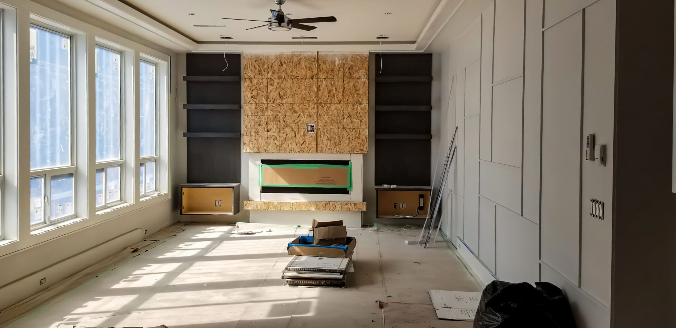 Living room under renovation with large windows, ceiling fan, and fireplace surrounded by built-in shelves, representing interior wall decoration in Quebec focused on modern home remodeling.