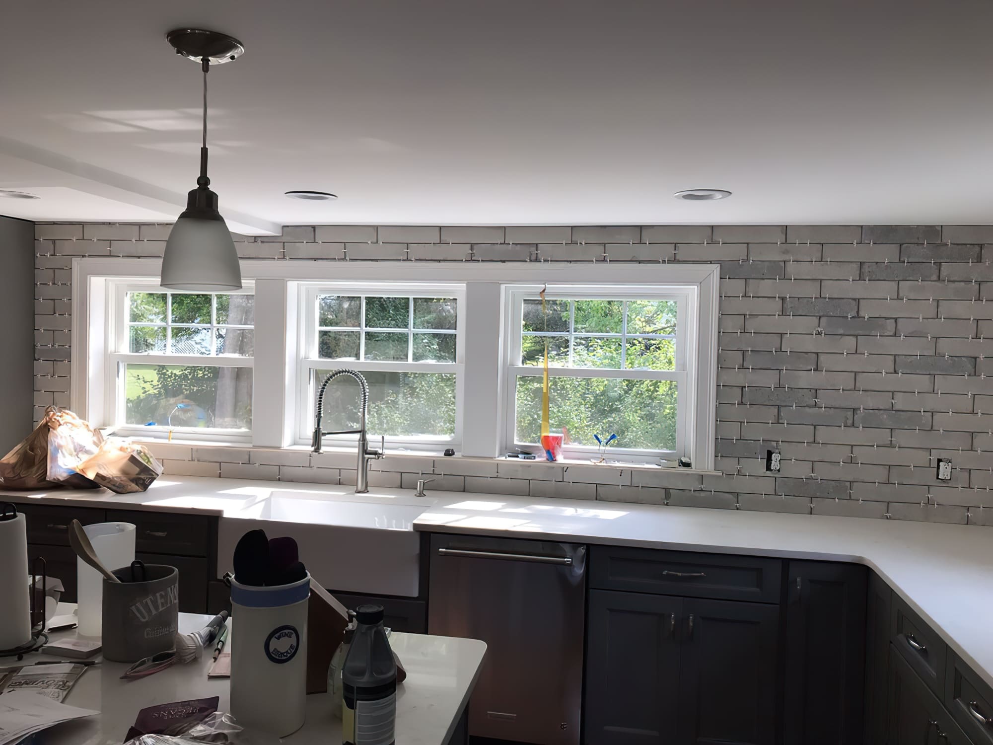 Residential renovations Quebec showing a kitchen under renovation with gray brick backsplash, farmhouse sink, white countertop, dark gray cabinets, and tools on the work surface.