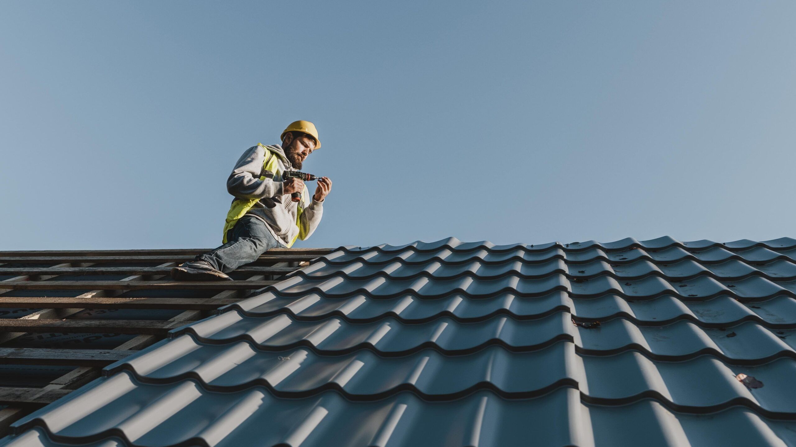 Roof replacement in Quebec showing a roofer wearing a yellow safety helmet and vest working on a flat metal-tile roof using a power tool, with details on proper slope and structural materials for flat roof construction.