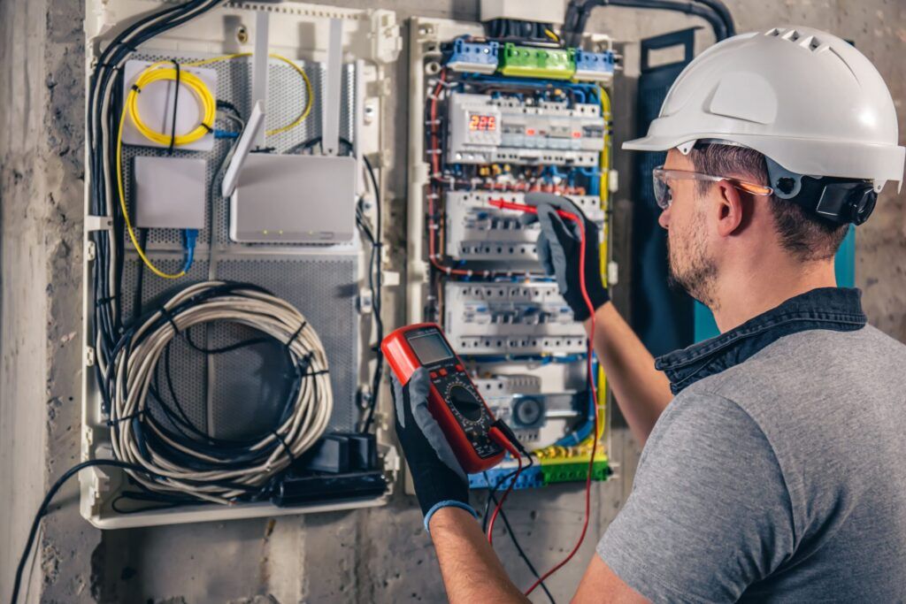 Electrician wearing safety gear using a multimeter to test a circuit breaker panel, representing professional mechanical systems services in Quebec focused on safe and efficient electrical maintenance.
