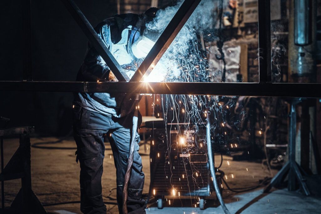 worker welding metal parts in a workshop Quebec — the image shows a metalworker using a welding torch to join metal components, with bright sparks visible; welding is a key process in metal fabrication involving heat and pressure to create durable joints used in construction, machinery, and manufacturing.