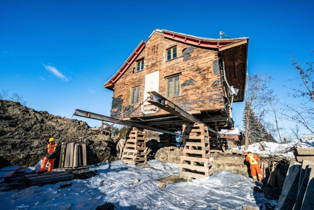 House relocation in Quebec showing a detached building supported by steel beams and temporary wooden structures while the ground is excavated below, with two workers in protective gear supervising the process for foundation replacement or site transfer.