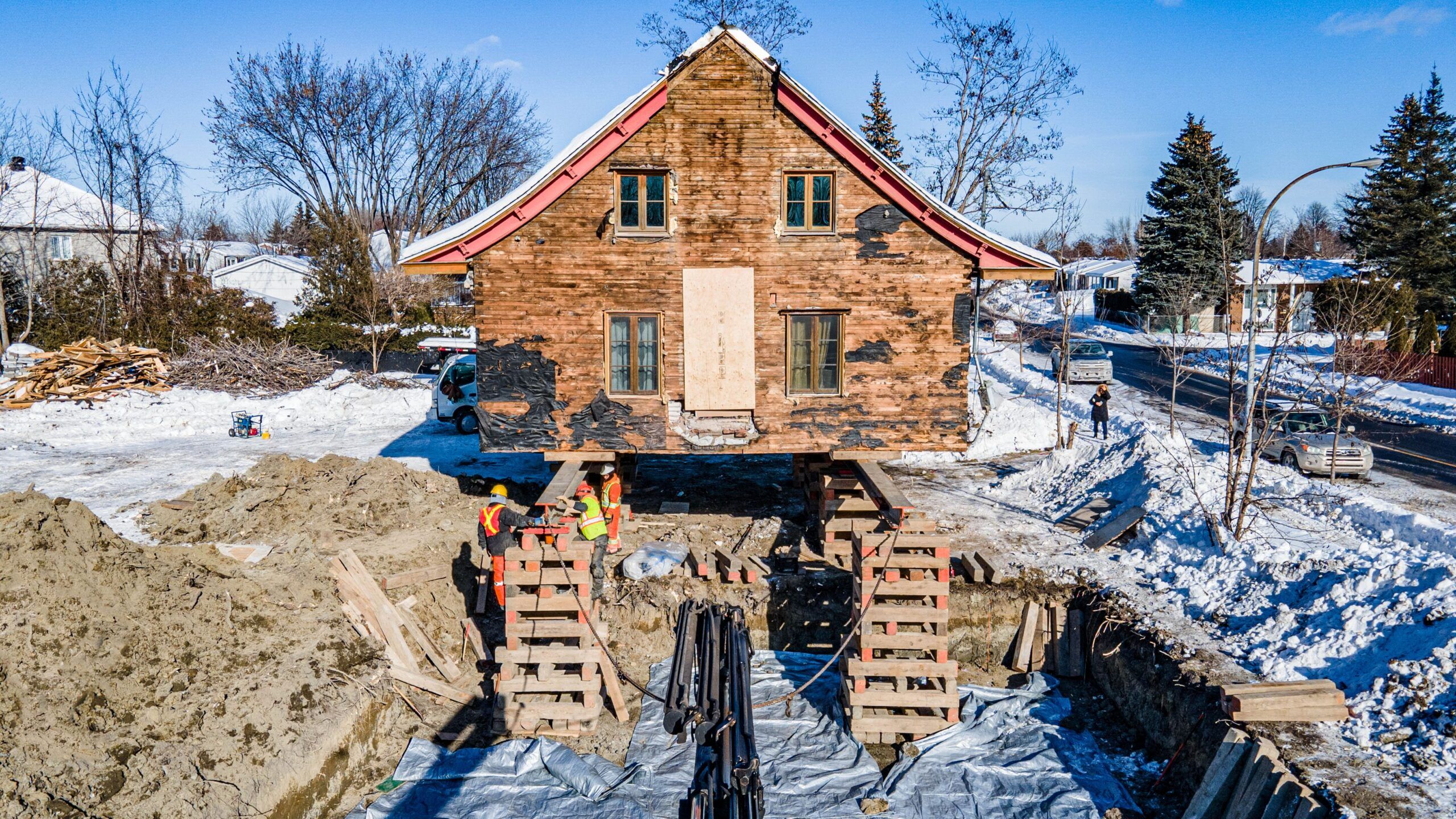 Lifting of Houses & Buildings Services Quebec showing a house elevated on steel beams and hydraulic jacks during relocation or foundation replacement, with workers preparing the new foundation in a snowy environment.