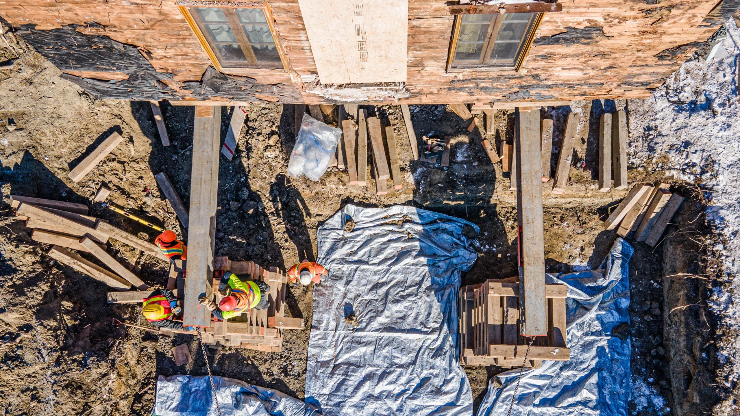 Lifting of Houses & Buildings Services Quebec showing an overhead view of a construction site where workers reinforce a brick building’s foundation with wooden and metal supports during foundation repair or stabilization work.