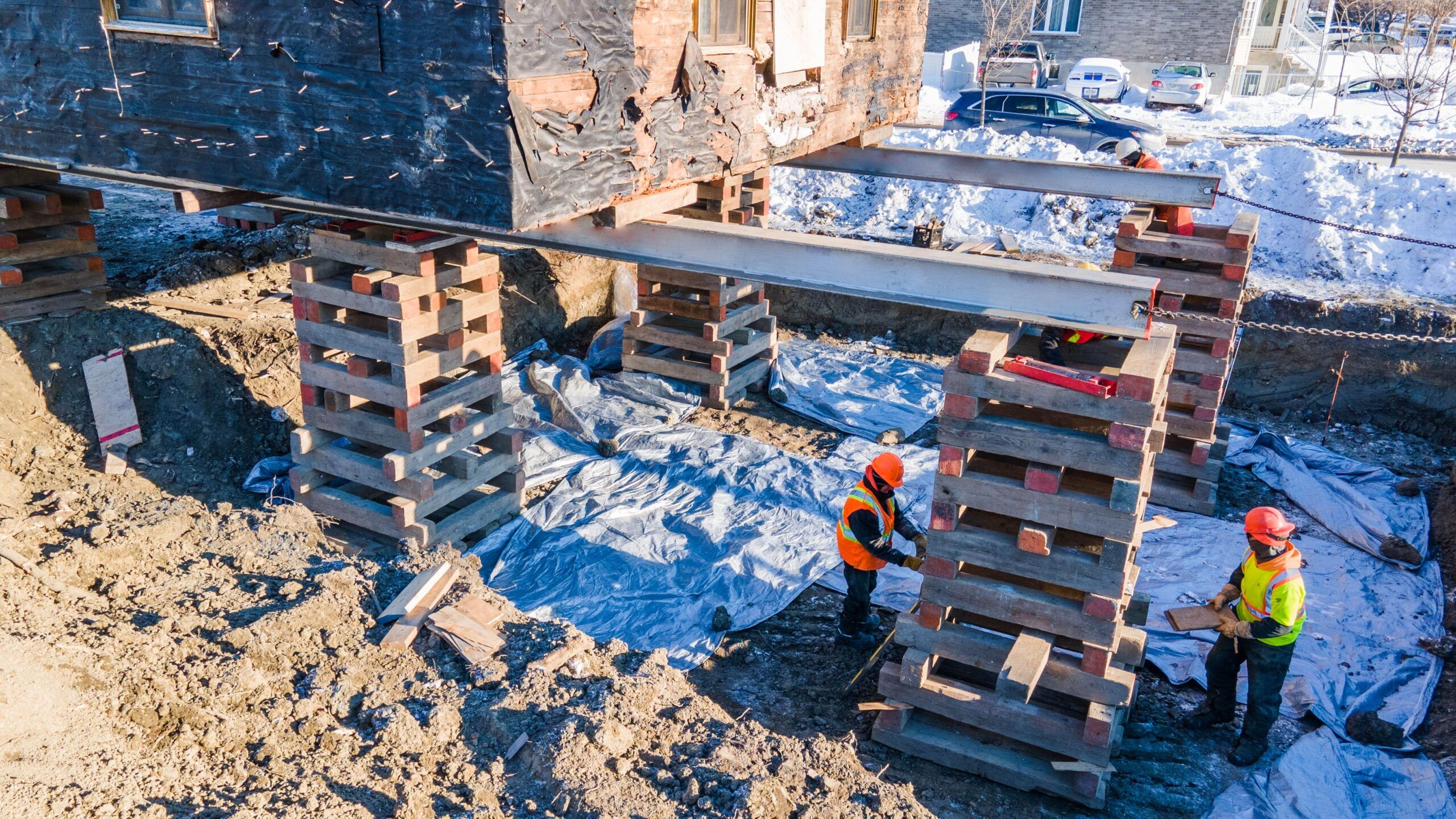 Lifting of Houses & Buildings Services Montreal showing a house elevated on steel beams and wooden supports for foundation repair or relocation, with workers in safety gear and snow-covered ground in the background.