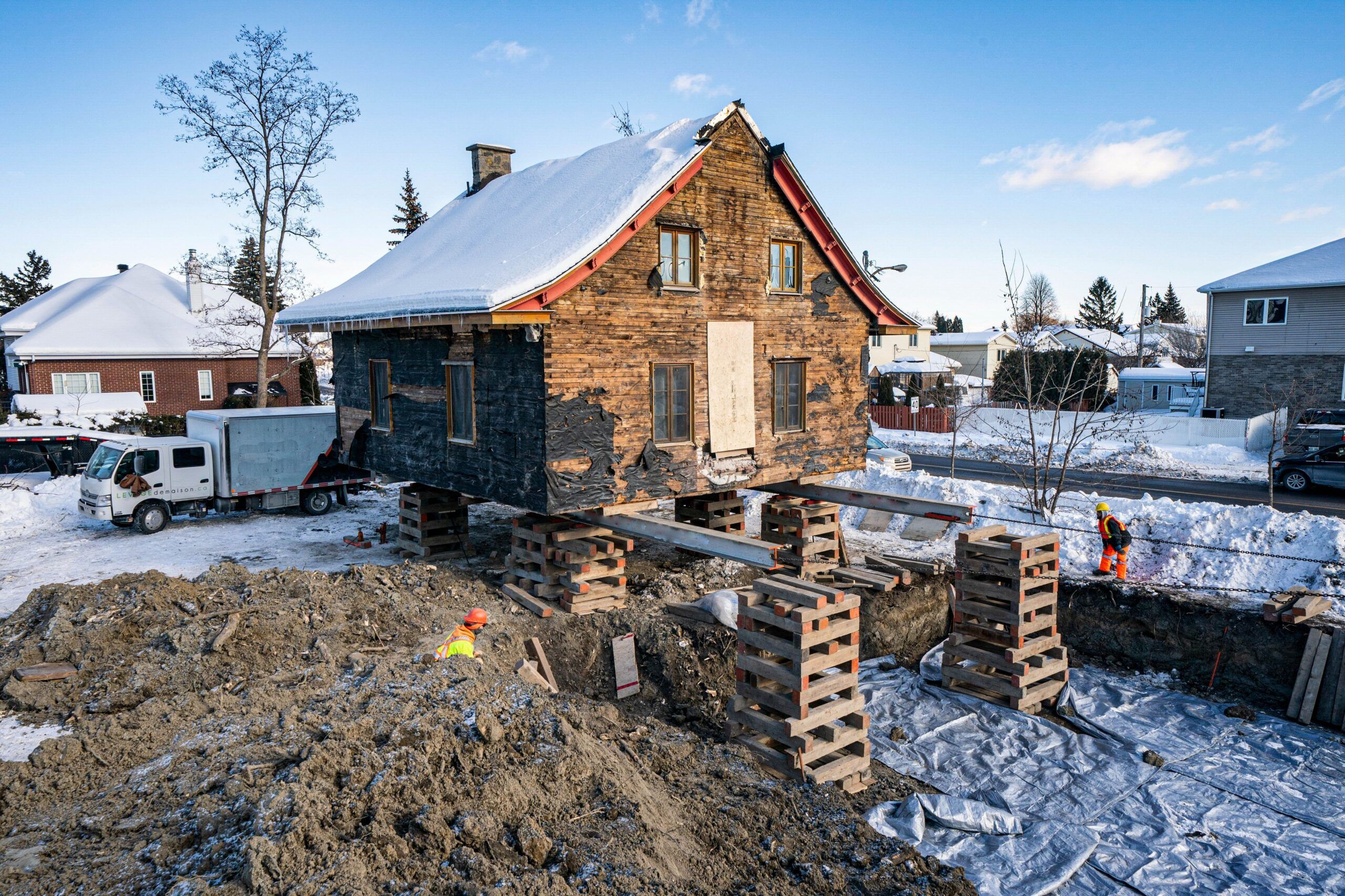 house lifting services Quebec and Montreal showing elevated home on wooden and steel beams in snowy environment with excavated foundation soil pile and two construction workers in orange safety vests.