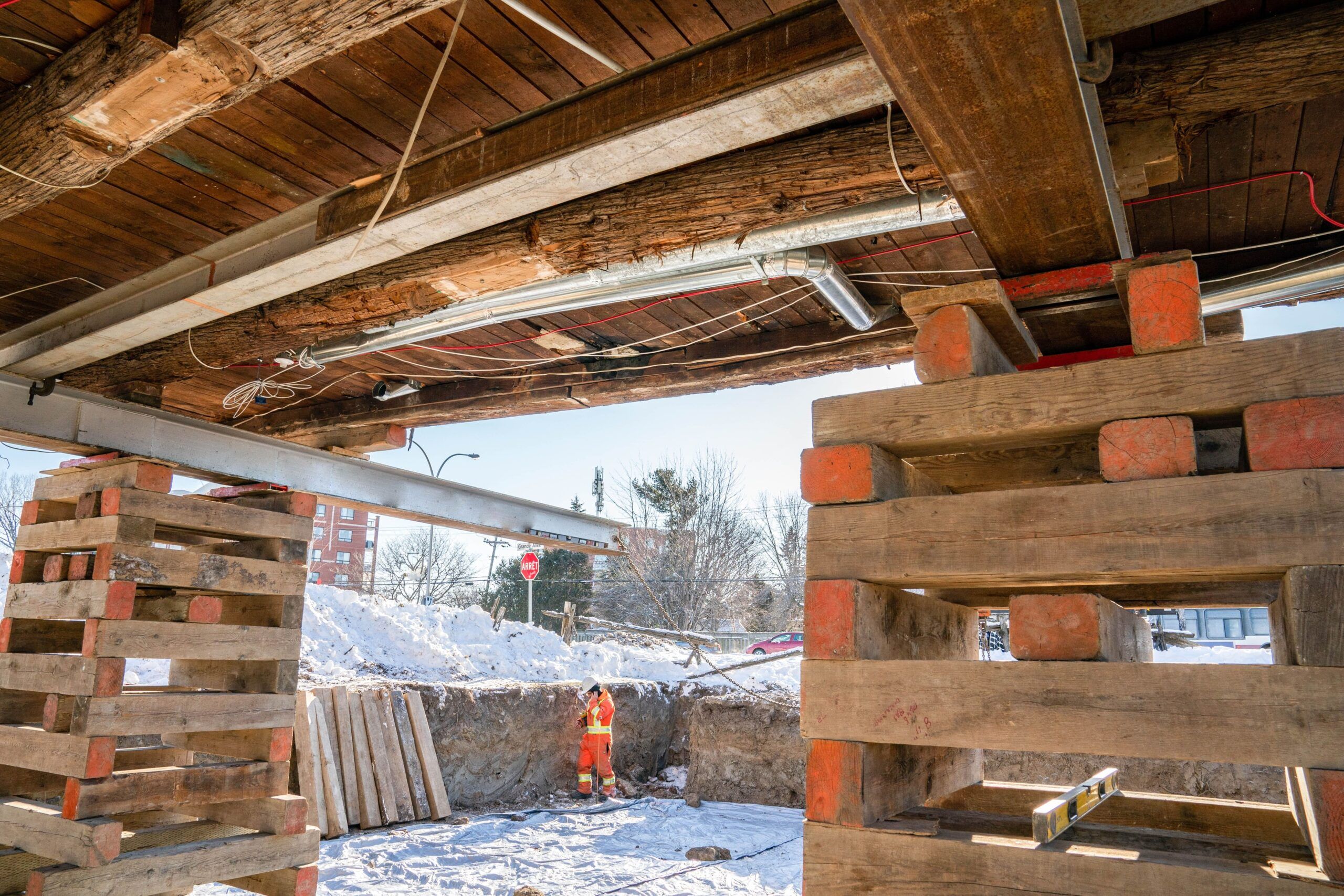 house lifting in Quebec with building supported on stacked wooden and steel beams, construction worker in orange safety gear working in snowy conditions, showing exposed wooden foundation structure during repair or relocation.